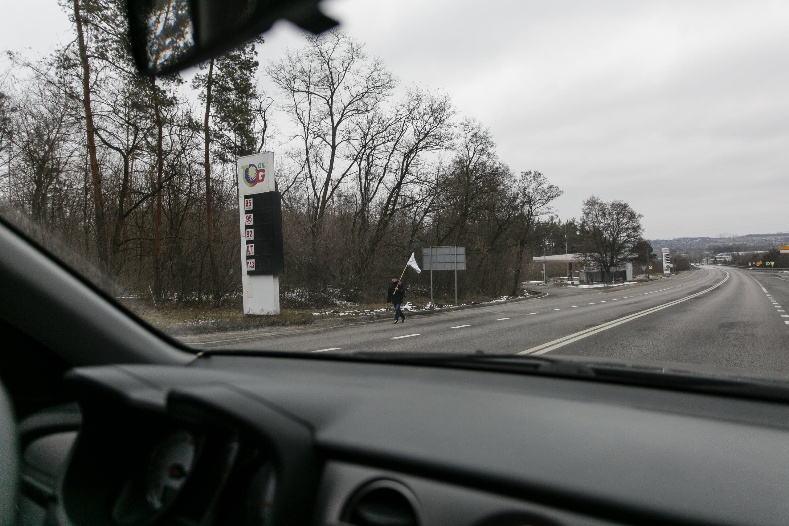 Вид из салона машины на трассу. На обочине, возле стелы АЗС ("OIL G", "95", "92", "ДТ", "ГАЗ"), идет человек с белым флагом.