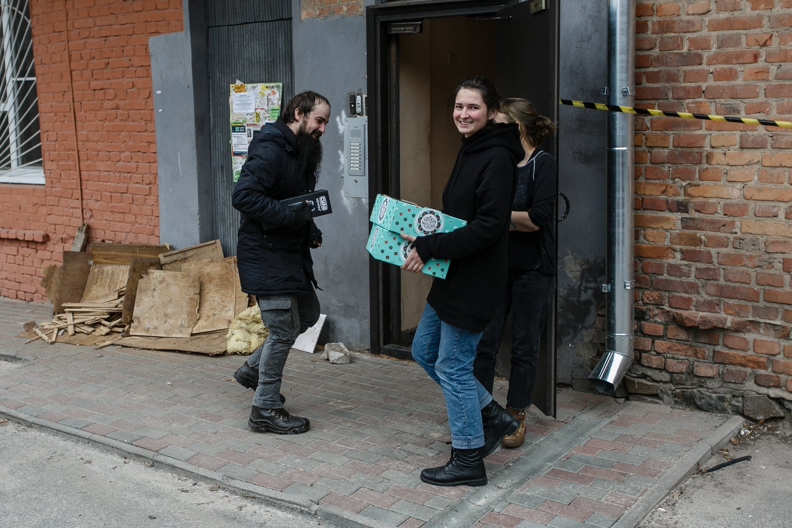 Женщина с бирюзовыми коробками и улыбающийся бородатый мужчина возле входа в кирпичное здание.