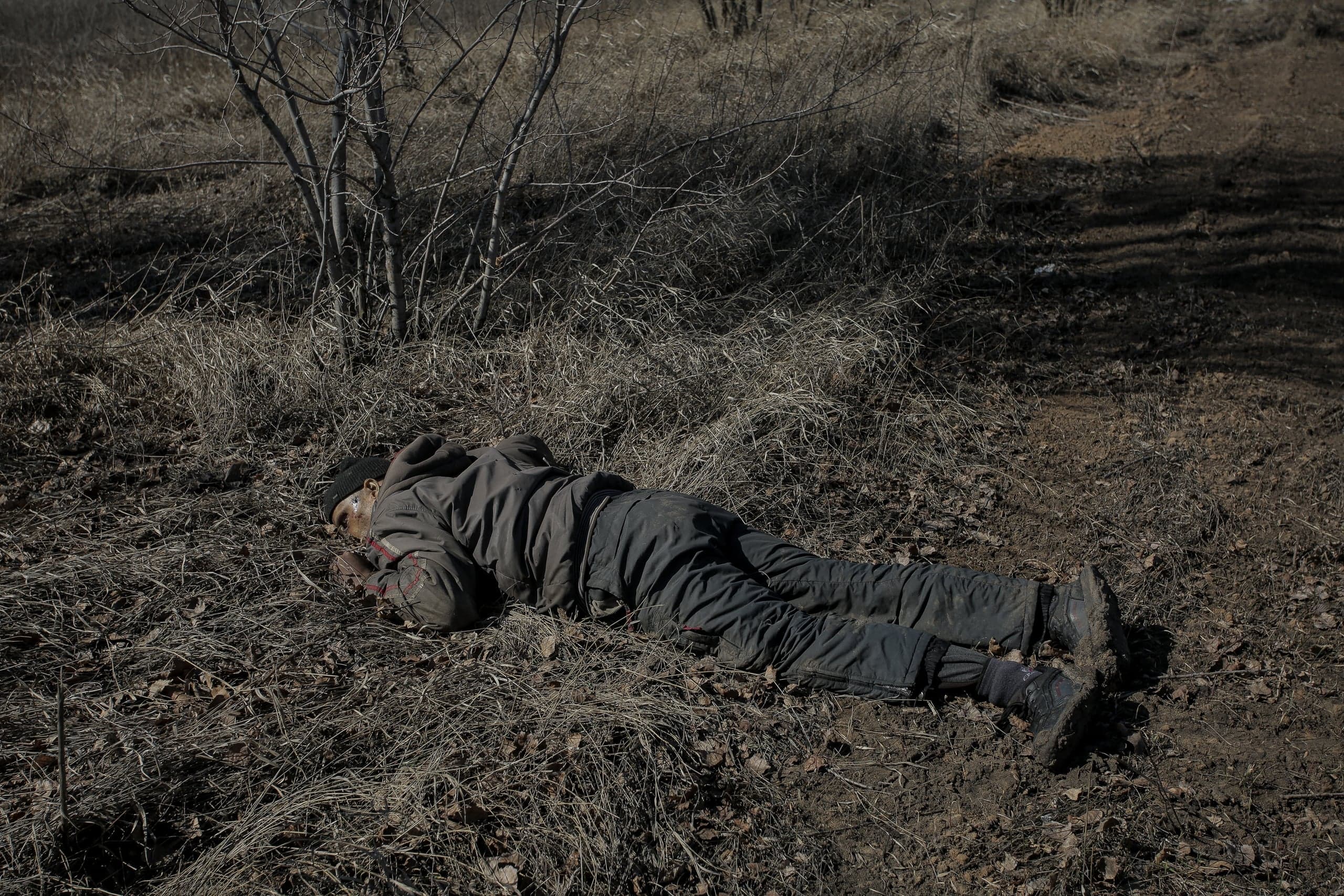 Тело человека в темной одежде, лежащее лицом вниз среди сухой травы и земли.