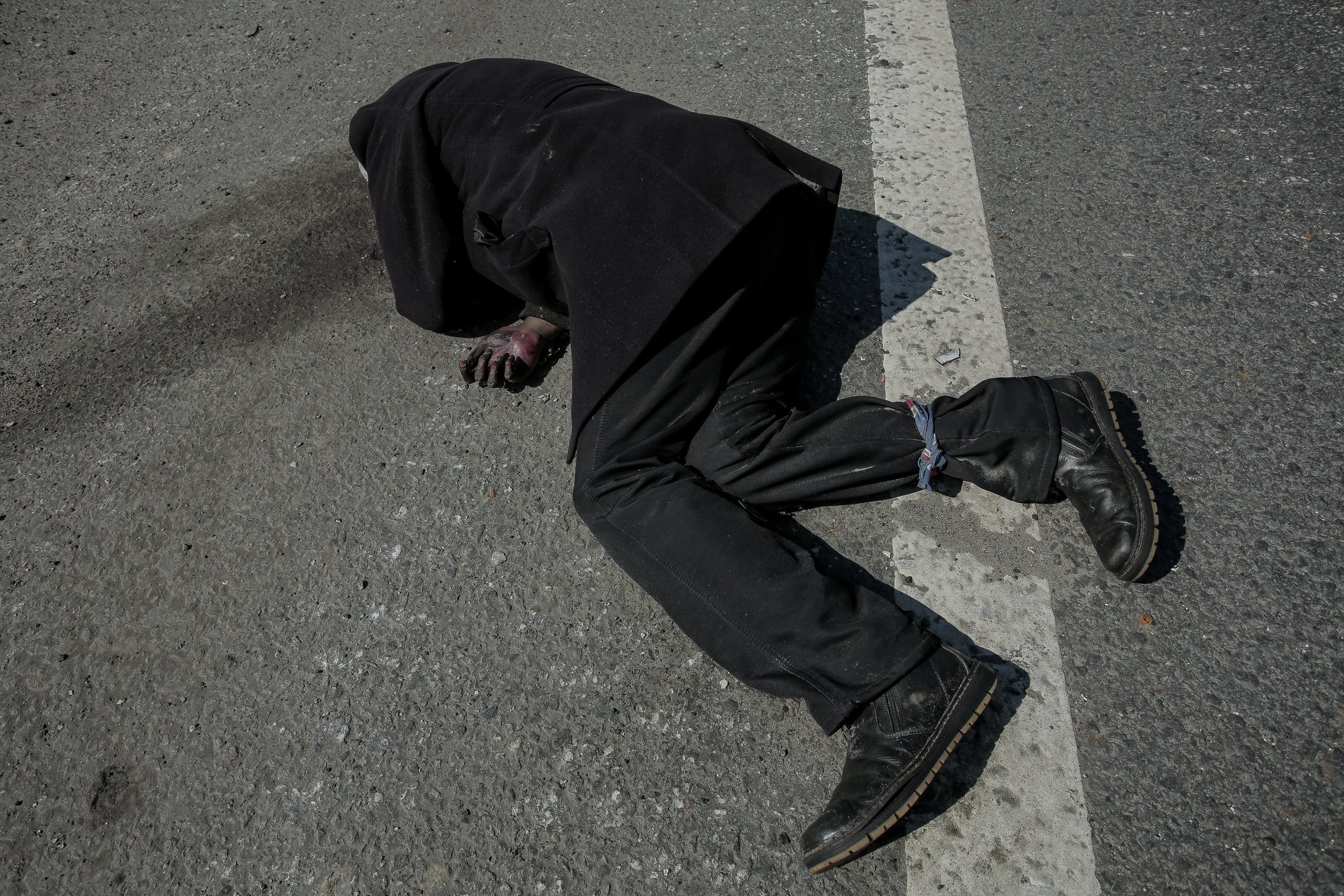Тело в черной одежде лежит лицом вниз на асфальте. Руки окровавлены, лодыжки связаны.