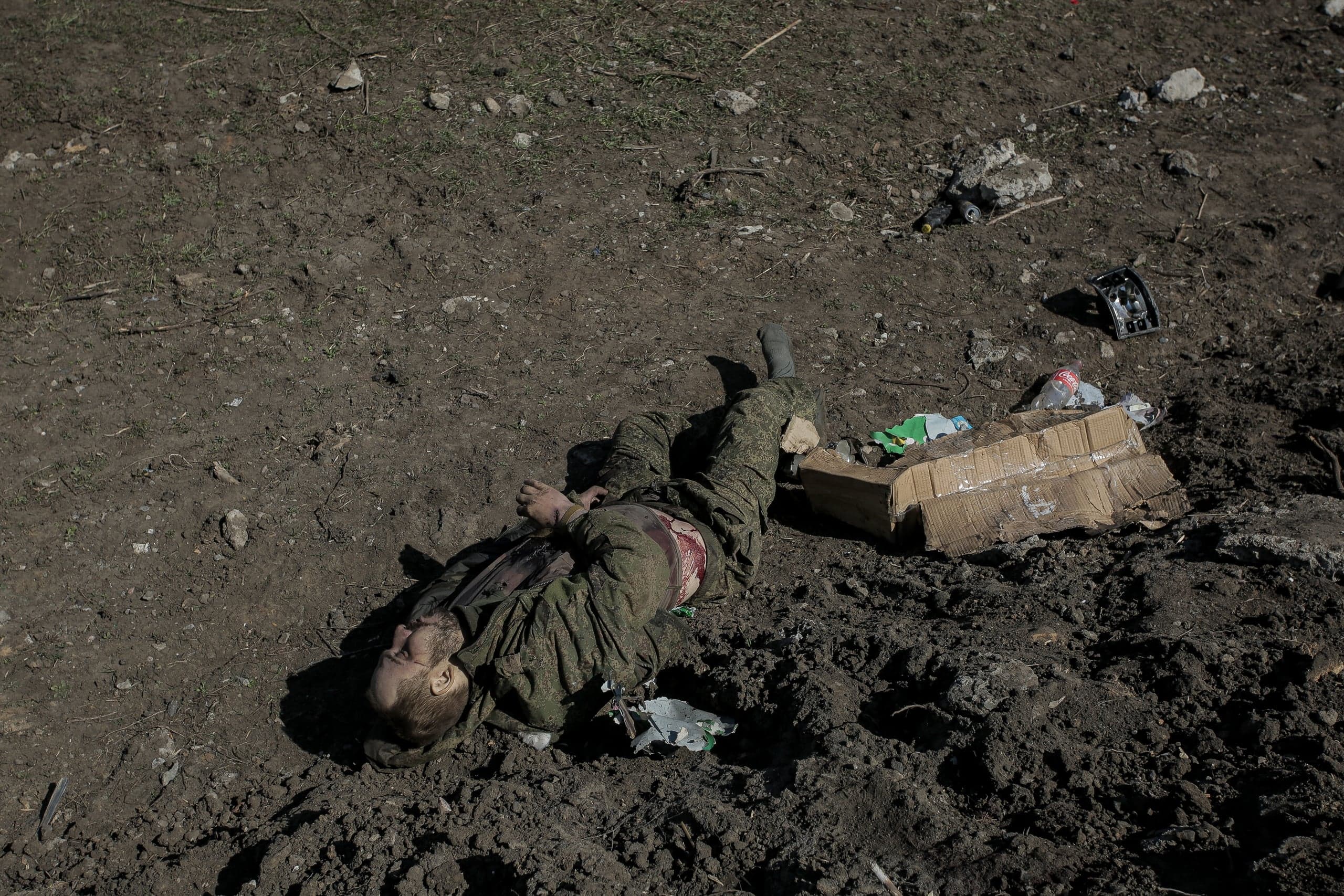 Тело военнослужащего в камуфляже с ранением живота лежит на грязной земле среди обломков.