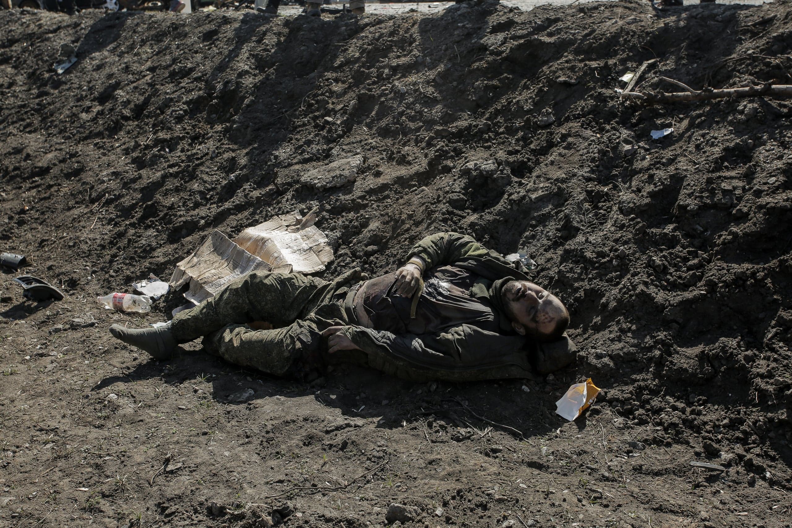 Тело солдата в камуфляже лежит на грязной земле у земляного вала. Вокруг разбросан мусор.