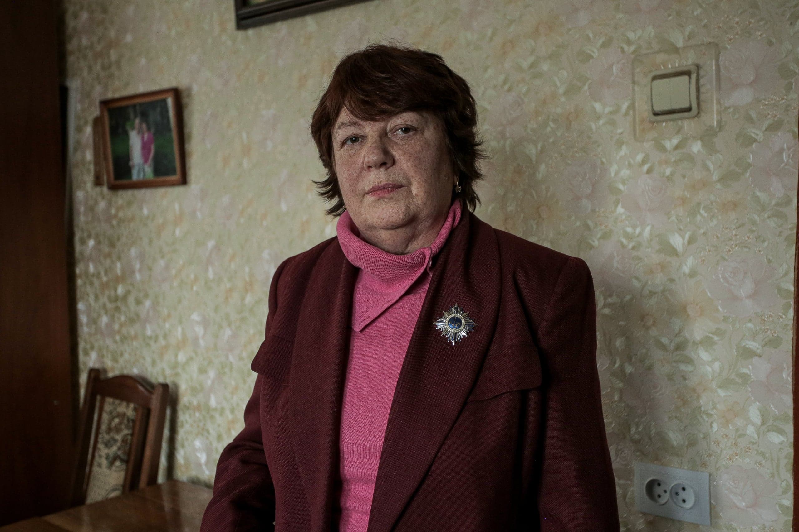 Портрет пожилой женщины в бордовом жакете с орденом на лацкане, стоящей на фоне светлых узорчатых обоев.