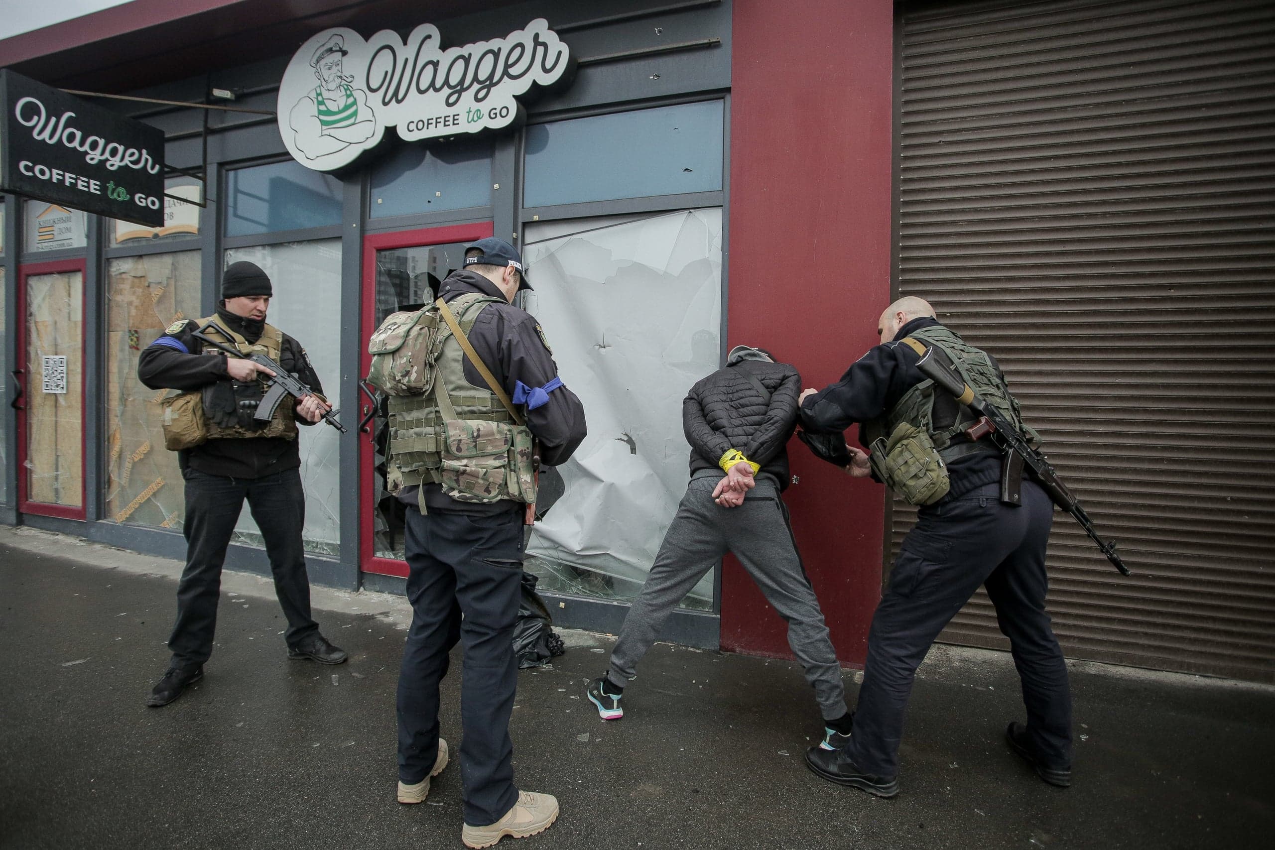 Трое вооруженных мужчин задерживают человека со связанными руками у поврежденного кафе. На вывеске: «Wagger COFFEE TO GO».