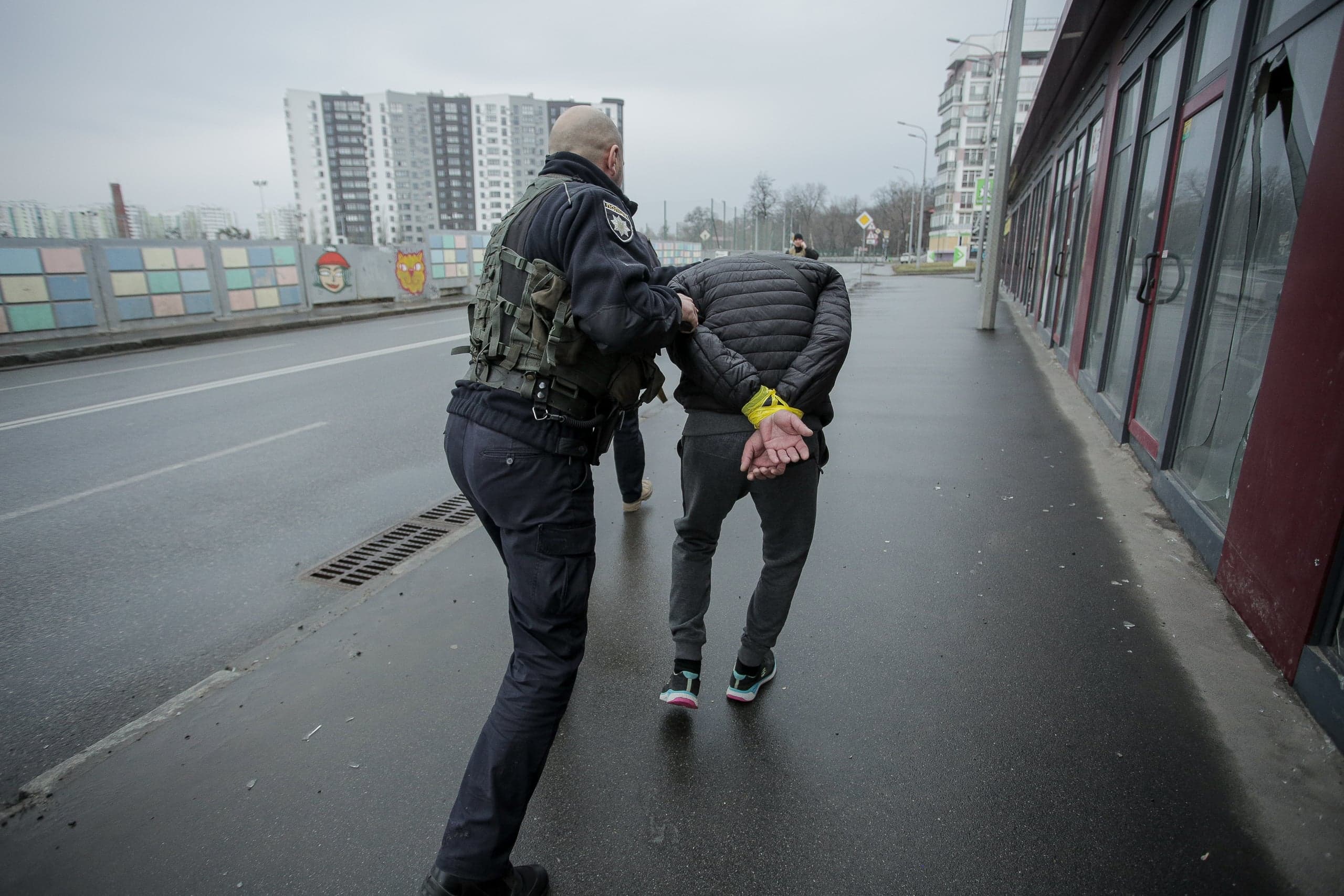 Офицер полиции ведет по мокрой улице мужчину с руками, связанными желтой лентой, мимо ряда зданий с разбитыми витринами.