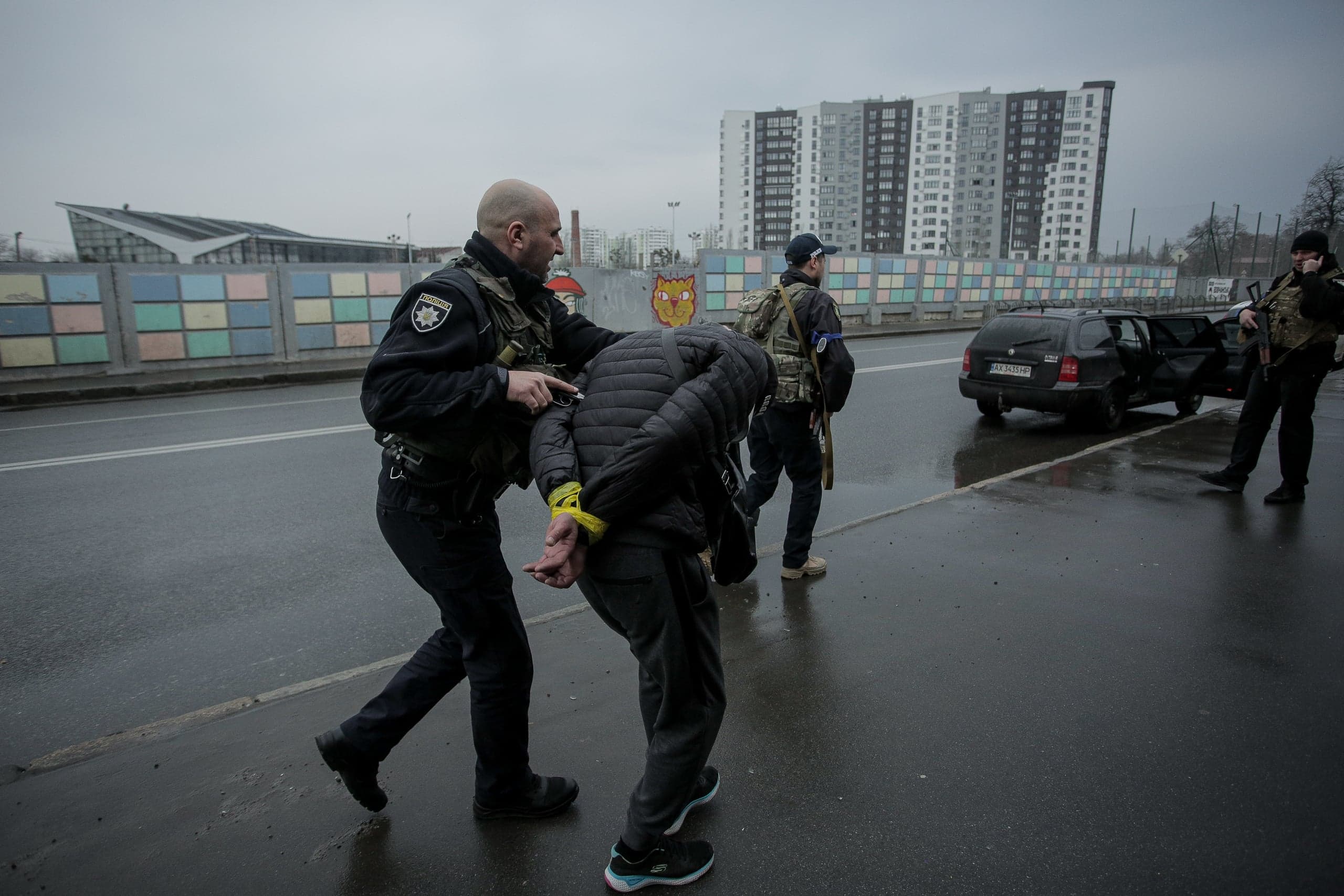 Украинский правоохранитель конвоирует задержанного, чьи руки связаны желтыми стяжками, по мокрой городской улице. На заднем плане — многоэтажные дома и служебный автомобиль.