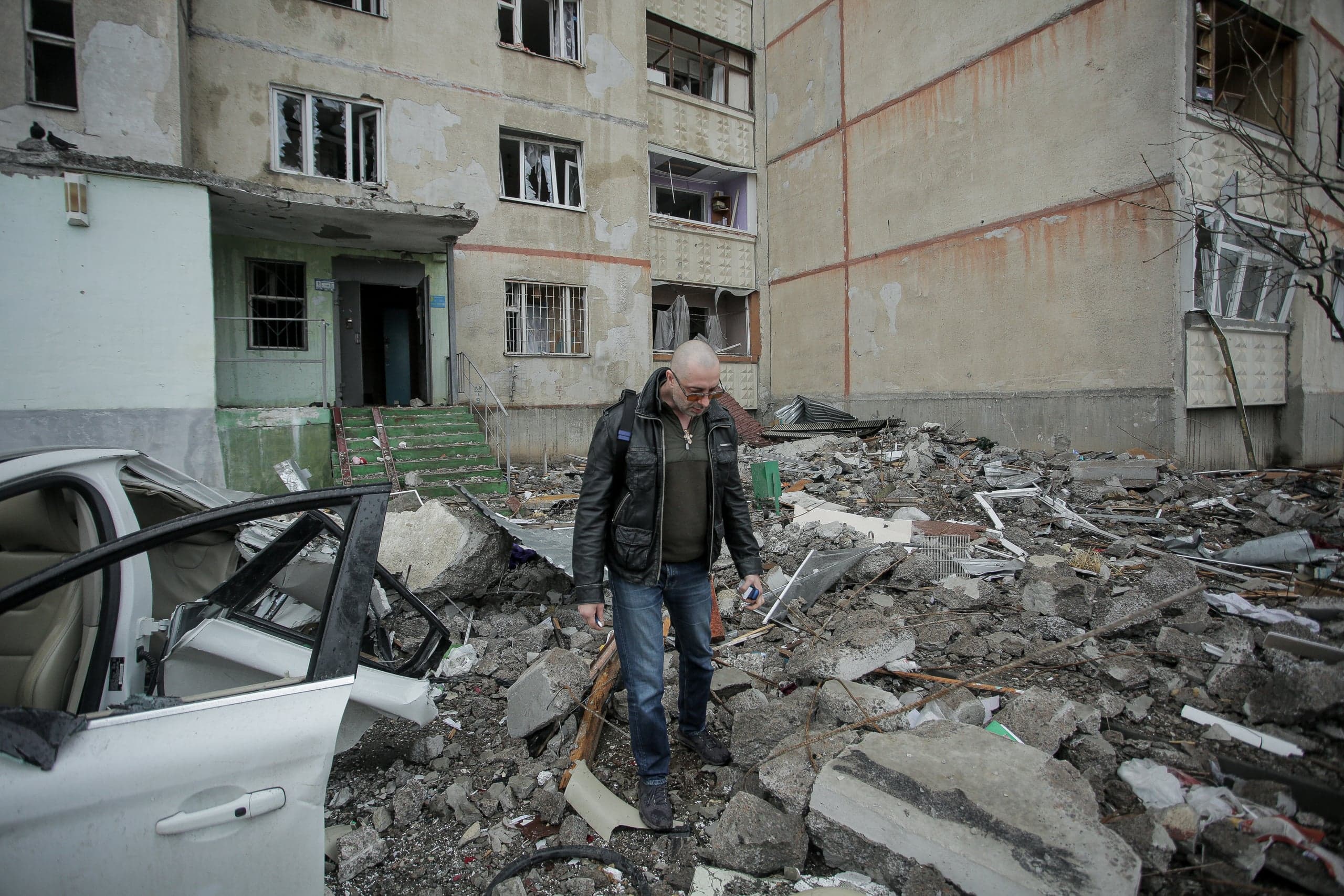 Мужчина идет по завалам и обломкам перед поврежденным многоквартирным домом; слева — разбитый автомобиль.