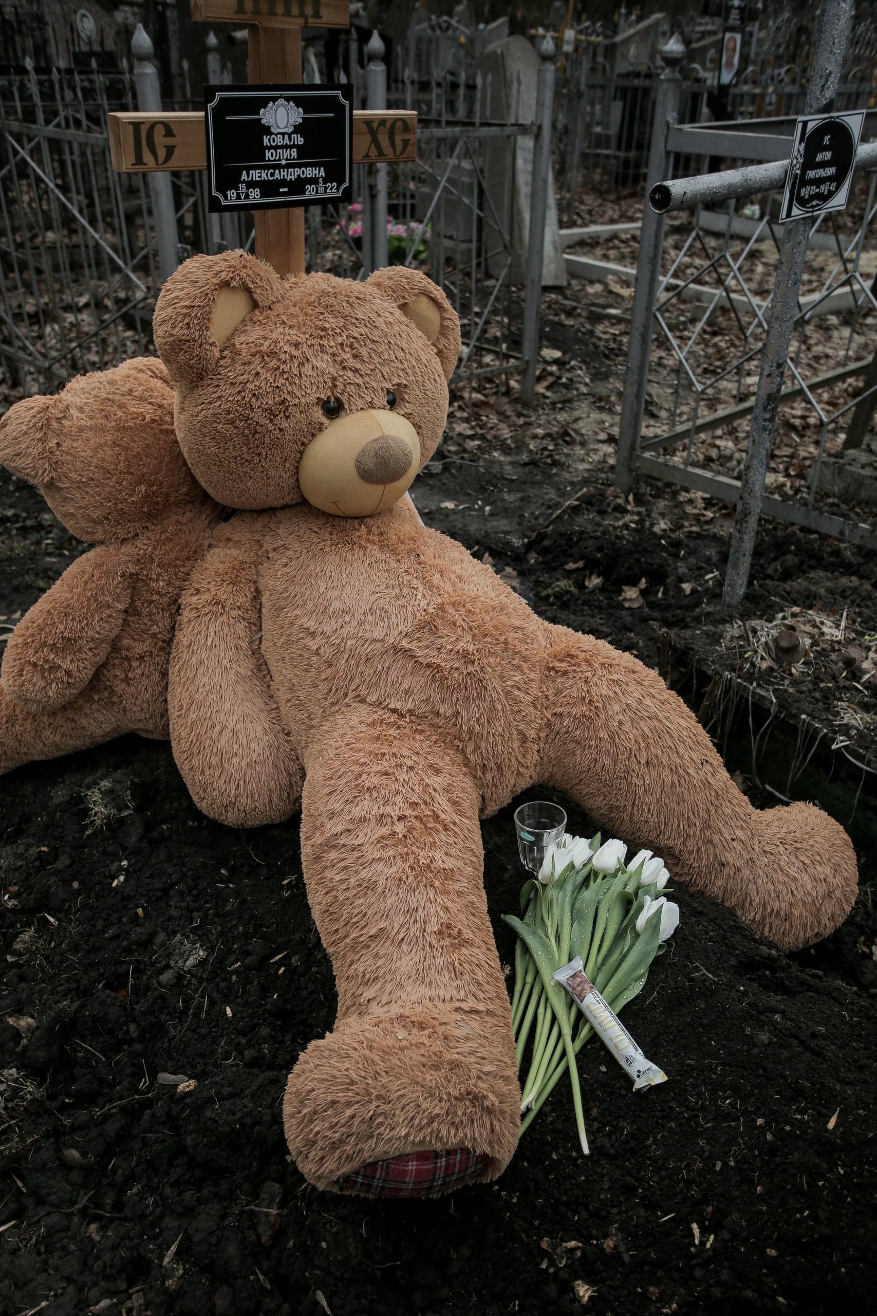 Плюшевый медведь, белые тюльпаны и стакан на могиле. На кресте видна табличка: "КОВАЛЬ ЮЛИЯ АЛЕКСАНДРОВНА 1998 – 2022".