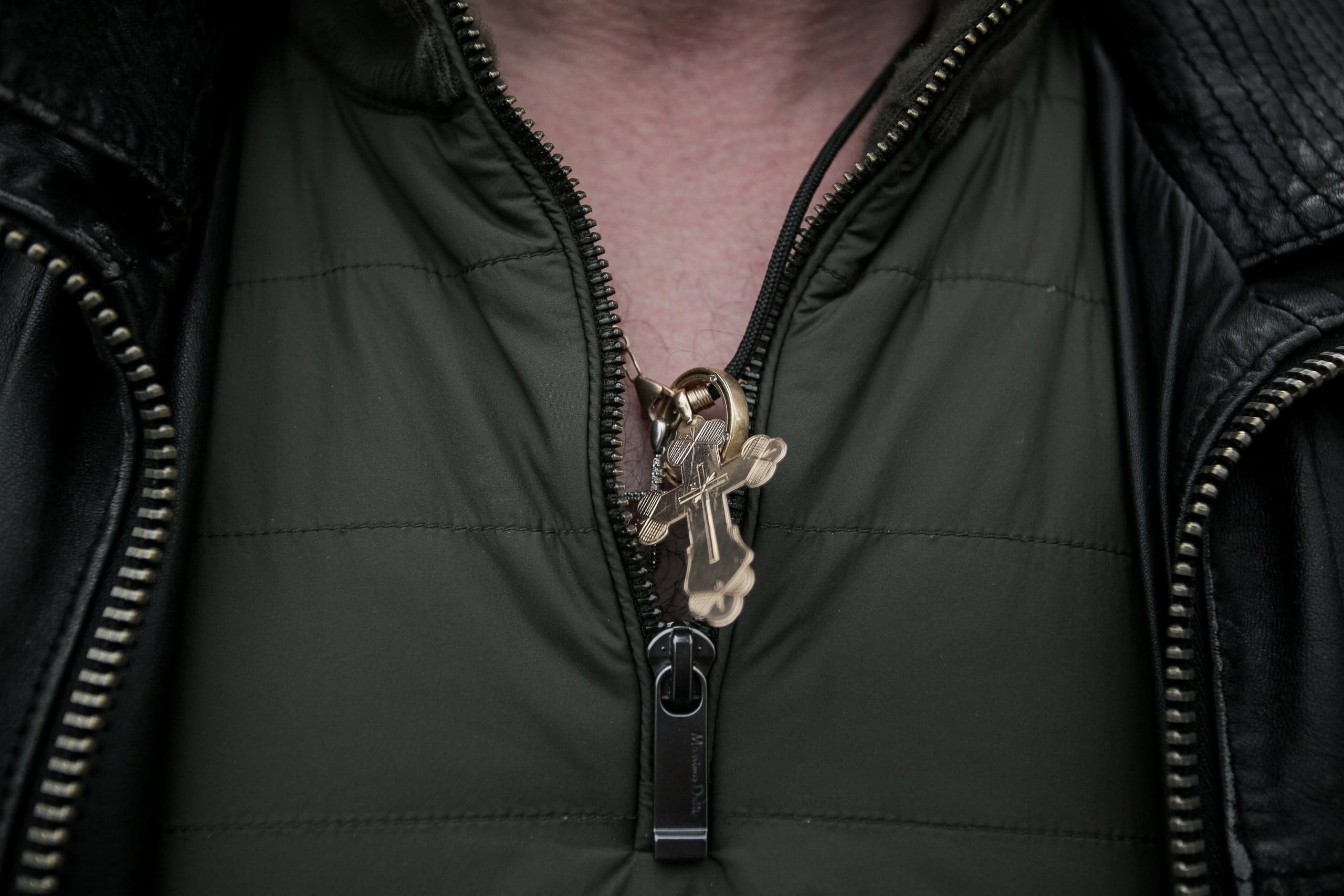 Золотой нательный крест, выглядывающий из расстегнутой оливковой куртки на фоне черной кожаной куртки.