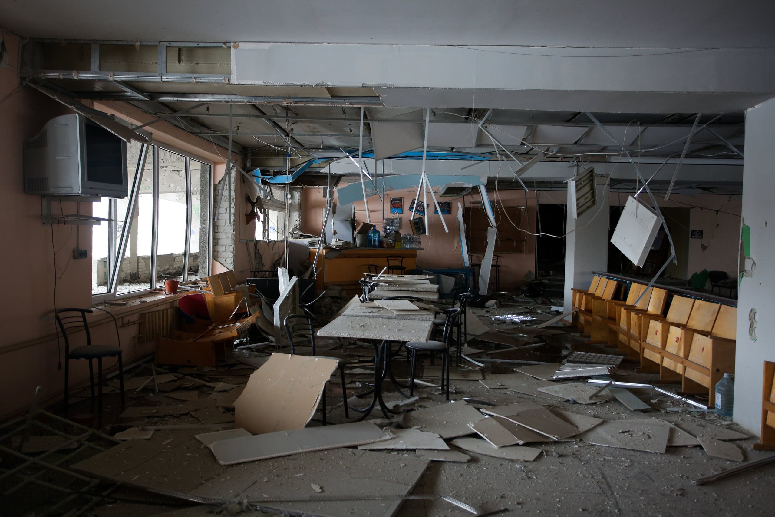 Интерьер сильно разрушенного помещения (возможно, кафе или зала): обрушенный подвесной потолок свисает на проводах, а пол покрыт грудой строительного мусора, обломками и поврежденной мебелью.