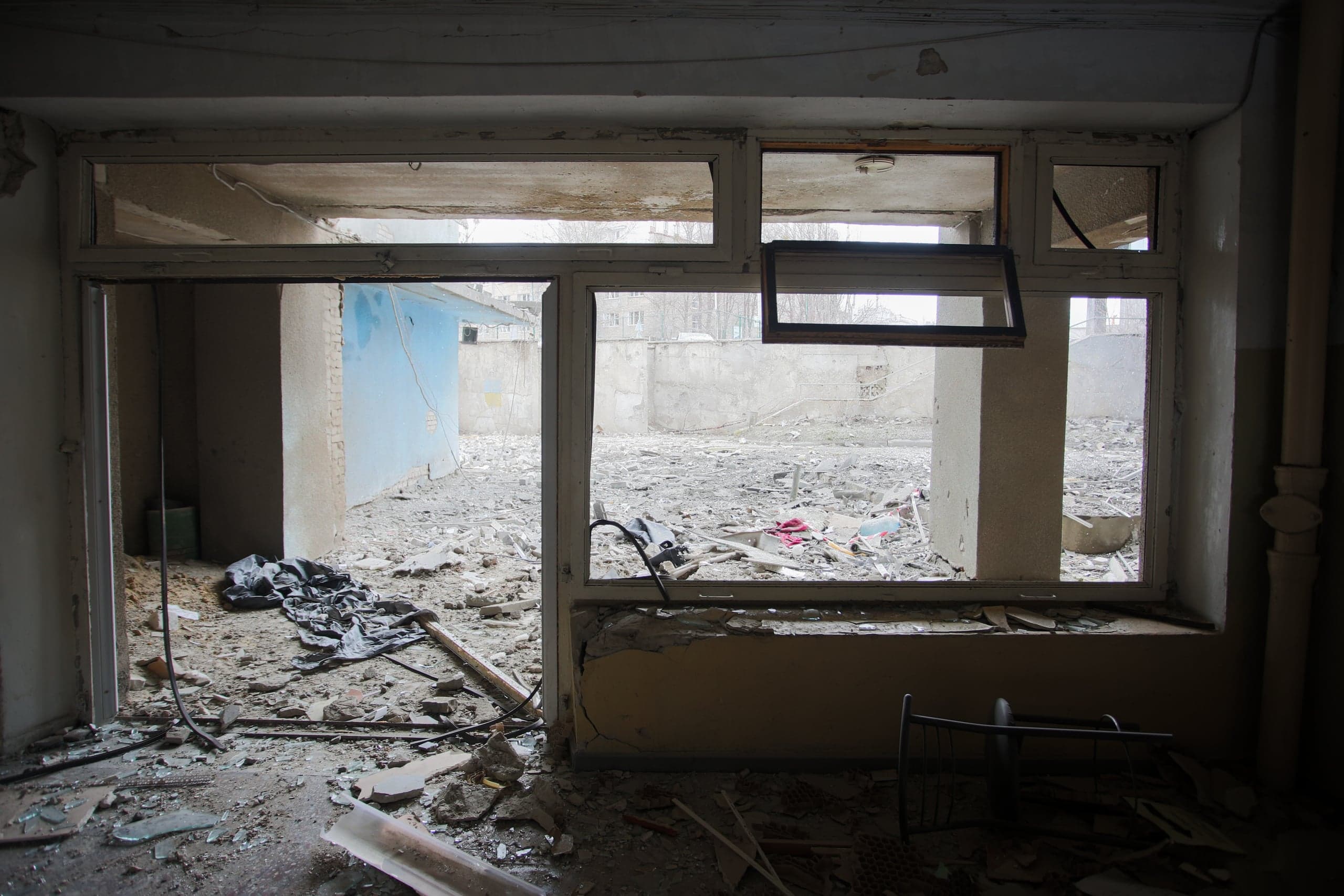 Вид из темного разрушенного помещения через пустой оконный проем на двор, заваленный строительным мусором и обломками.