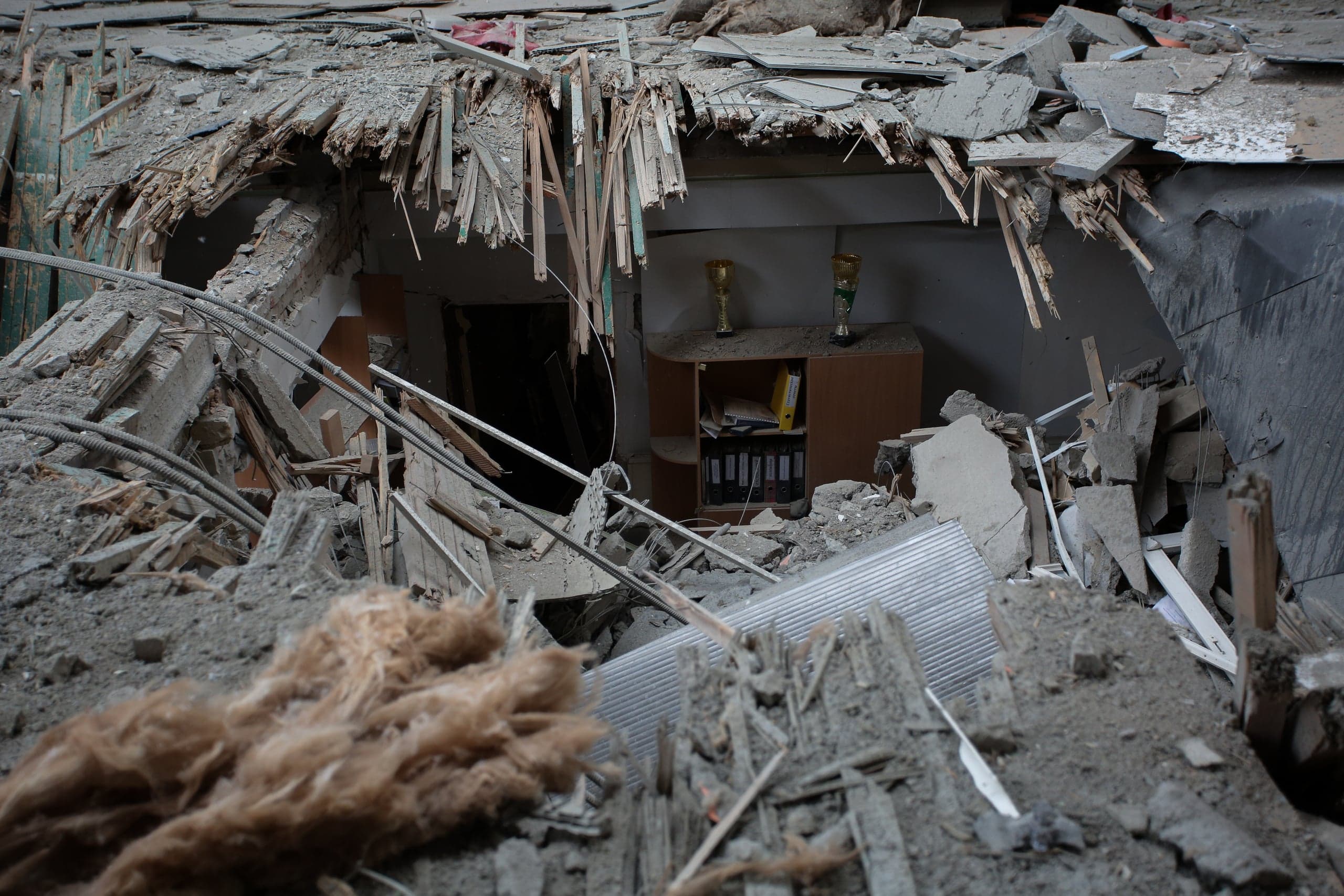 Крупный план разрушенного помещения: груды бетона, обломков и арматуры перед проломом. Через пролом виден пыльный офисный шкаф с папками и двумя кубками (трофеями).