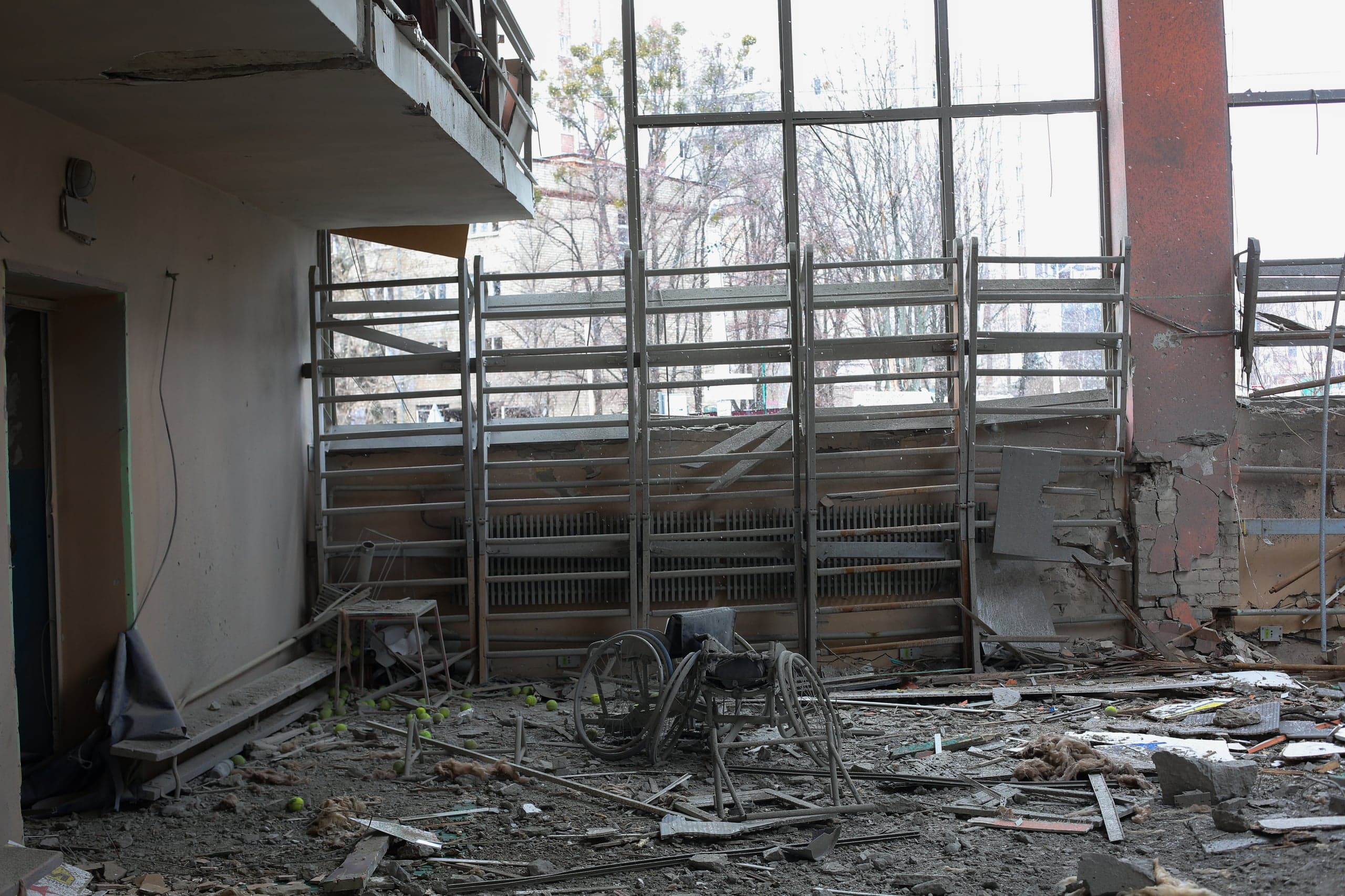 Разрушенный интерьер спортивного зала: пол покрыт обломками и мусором, среди которых лежат сломанное инвалидное кресло и множество теннисных мячей. На заднем плане видны большие окна и поврежденные гимнастические стенки.