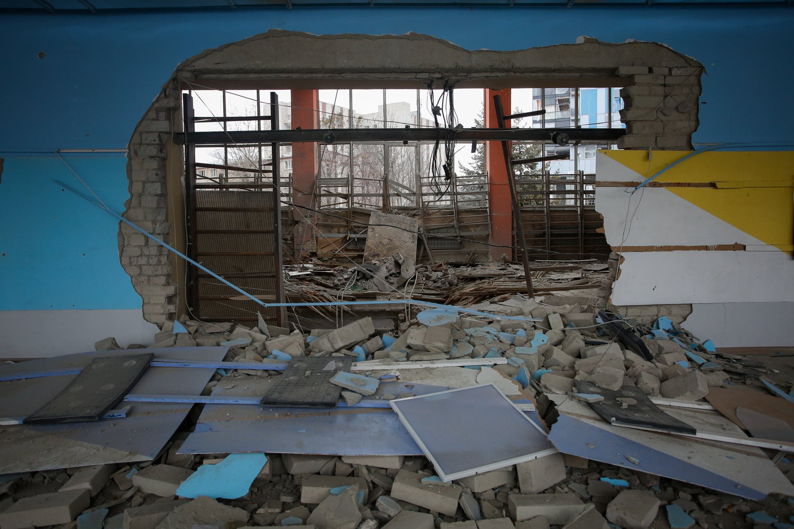 Интерьер сильно поврежденного здания с большой рваной дырой в стене (окрашенной в синий и желтый цвета). Перед проемом лежит груда строительного мусора и обломков, а за ним видны металлические каркасы и многоэтажные дома на фоне.