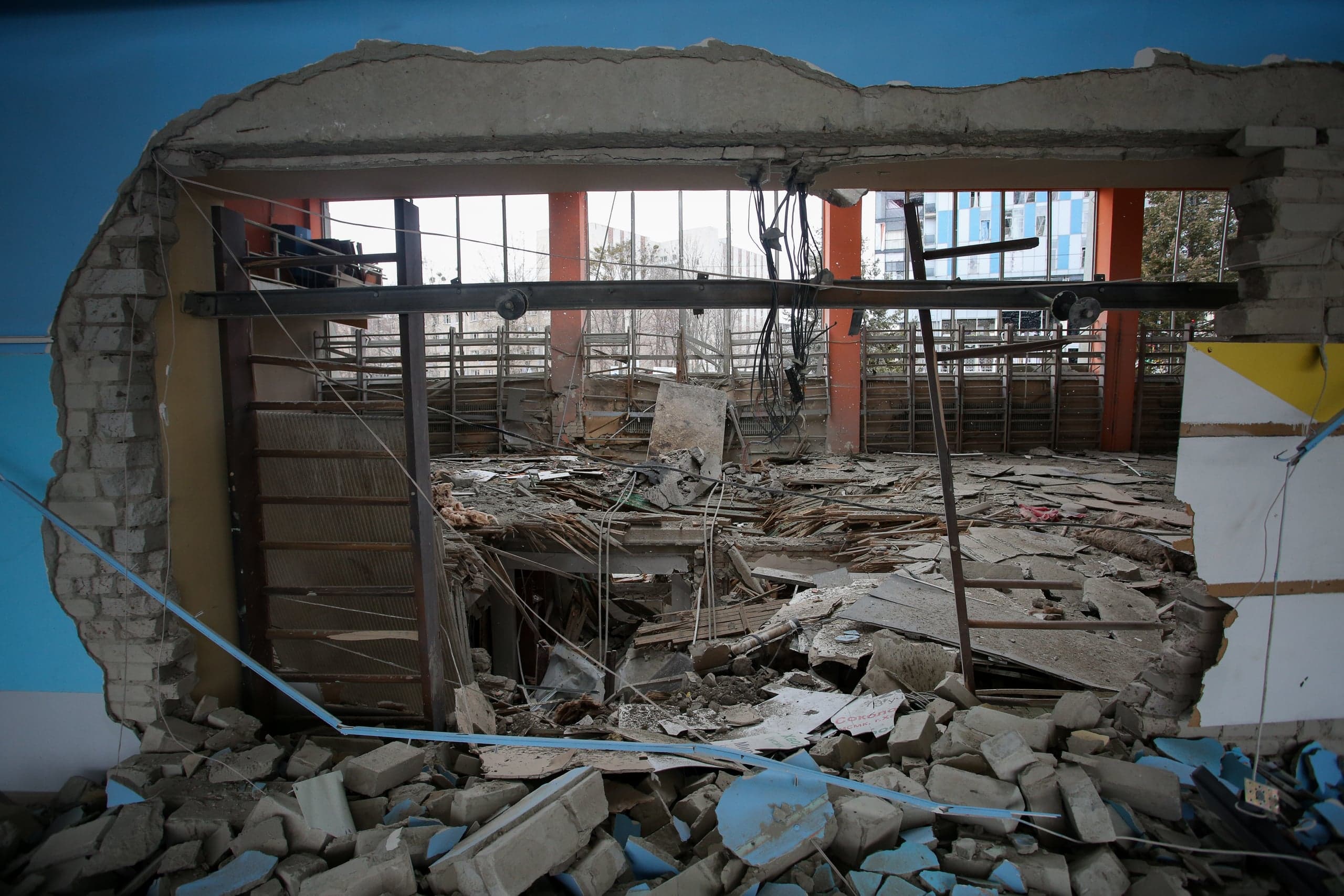 Крупный план проема в синей стене, заваленного грудой строительного мусора и обломков. Через проем видно полностью разрушенное внутреннее помещение с грудами досок, подвешенными проводами и оранжевыми структурными колоннами.