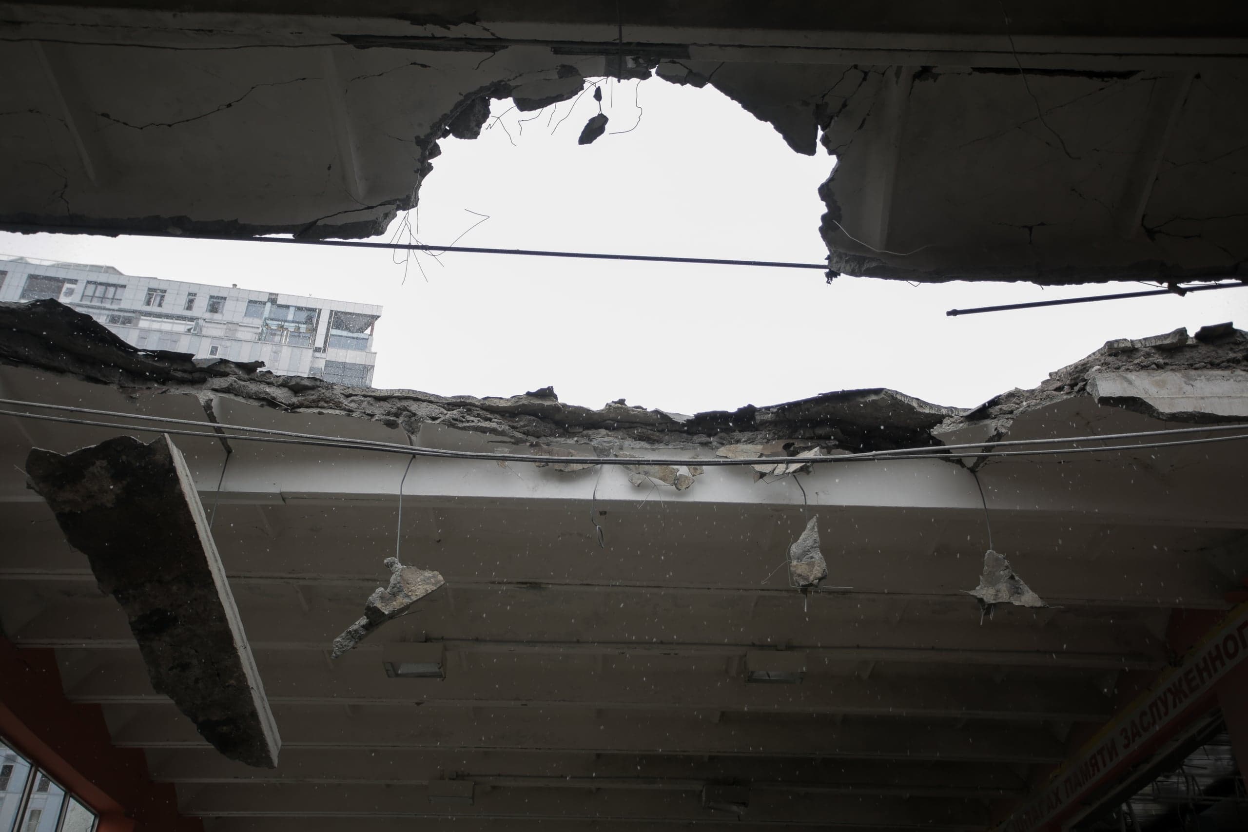 Вид снизу на огромное рваное отверстие в разрушенном бетонном перекрытии. Свисают обломки и арматура. Сквозь проем видны светлое небо и часть городского здания на заднем плане.