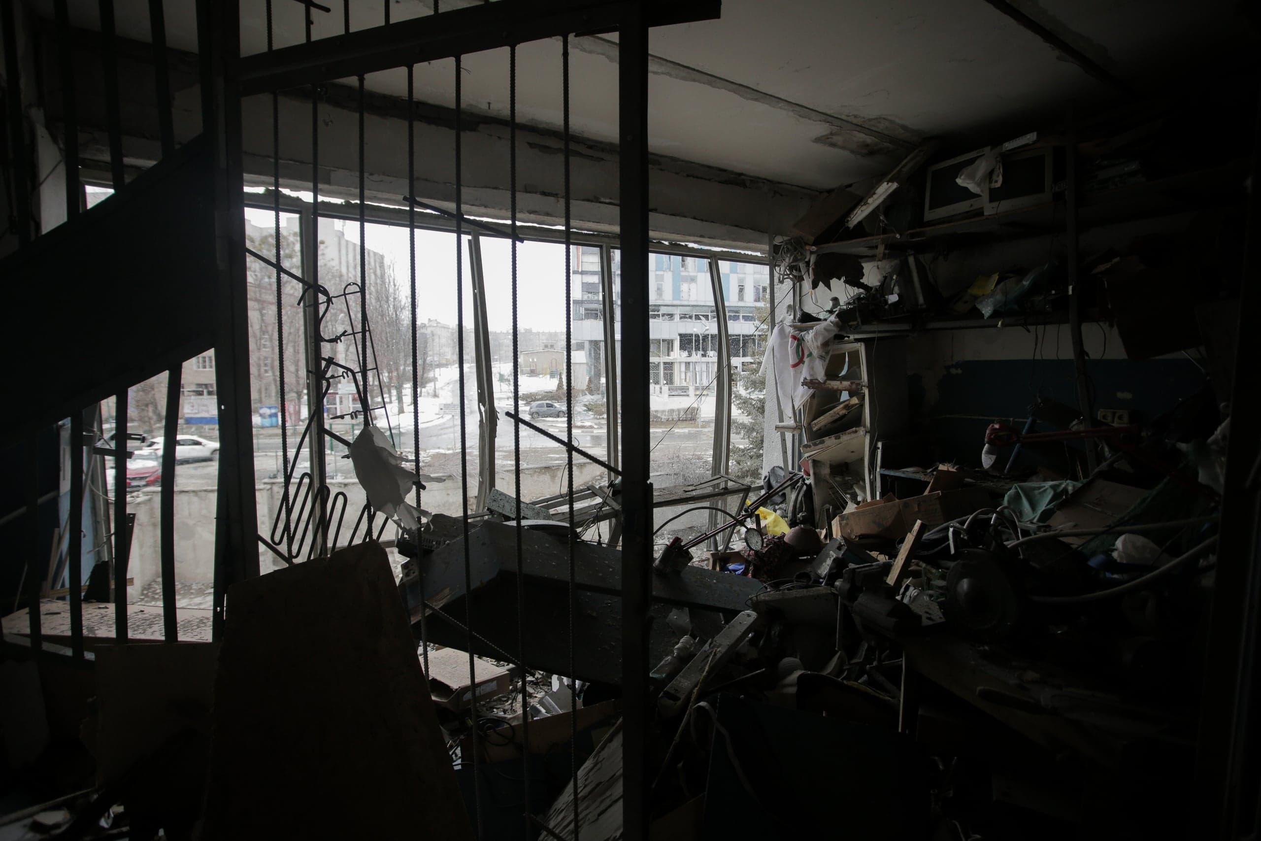 Силуэтный снимок изнутри разрушенного здания: комната завалена обломками и мусором, сквозь разбитый оконный проем с металлическими прутьями видно заснеженную городскую улицу.