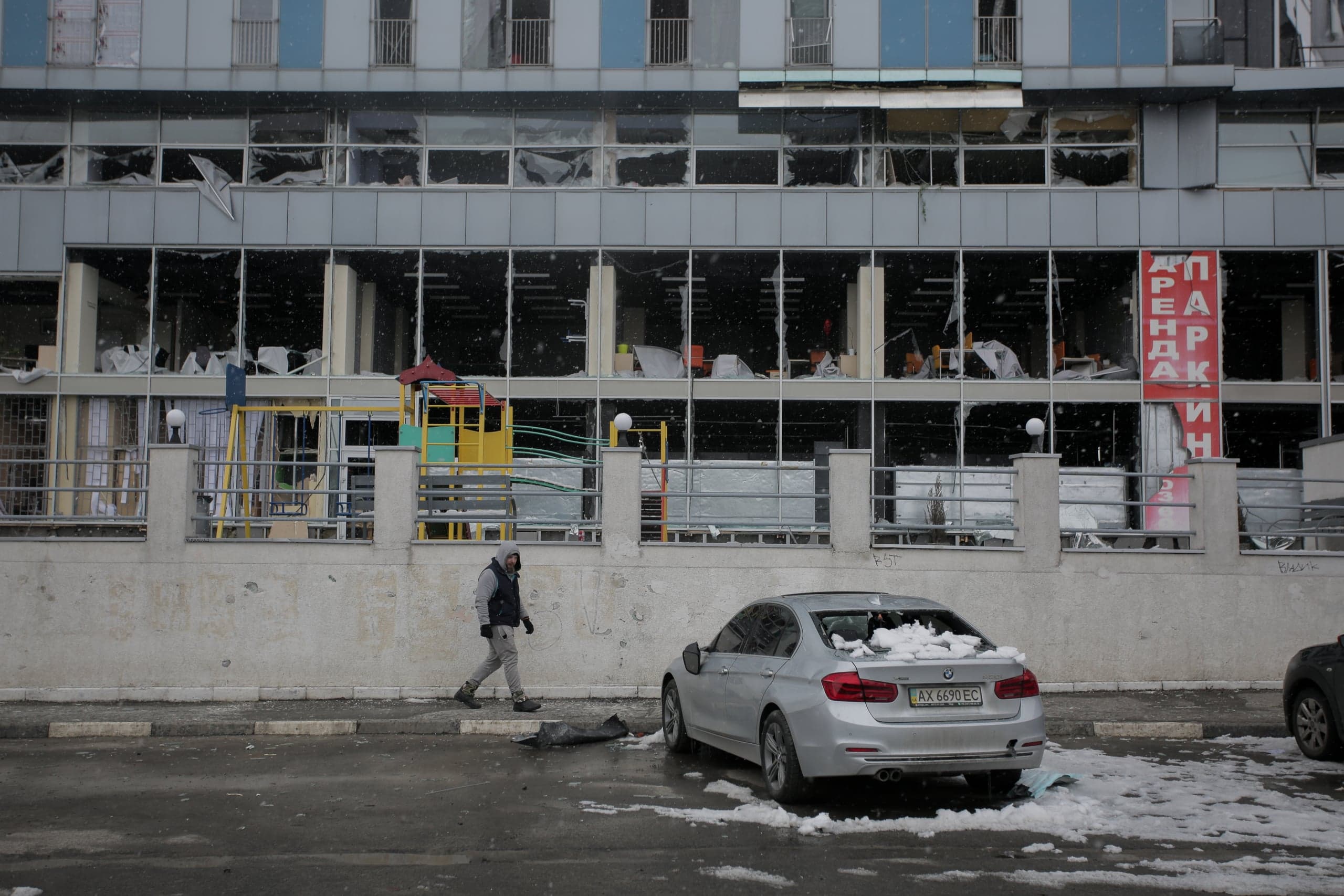 Здание с многочисленными разбитыми окнами на фасаде. На переднем плане видна припаркованная серебристая машина с выбитым задним стеклом и проходящий мимо человек во время небольшого снегопада.