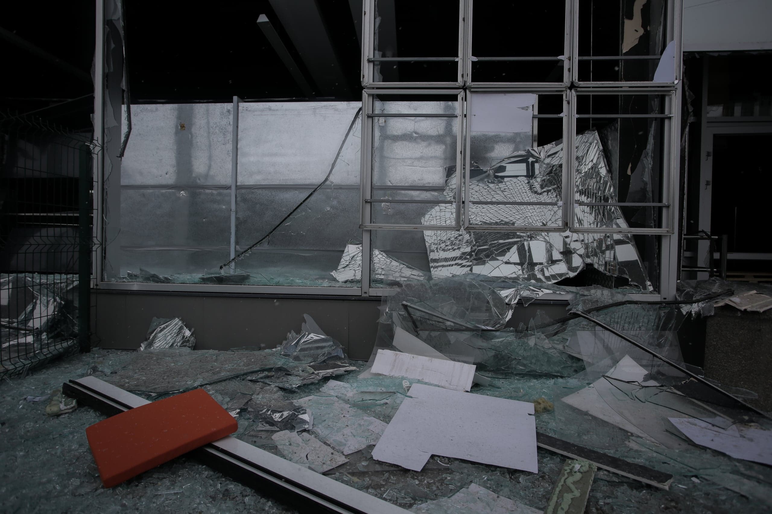 Крупный план разбитого стеклянного фасада здания. Наружные оконные рамы повреждены, а земля перед ними полностью усеяна грудой крупных и мелких осколков стекла и обломков. Слева внизу лежит оранжевый прямоугольный предмет.