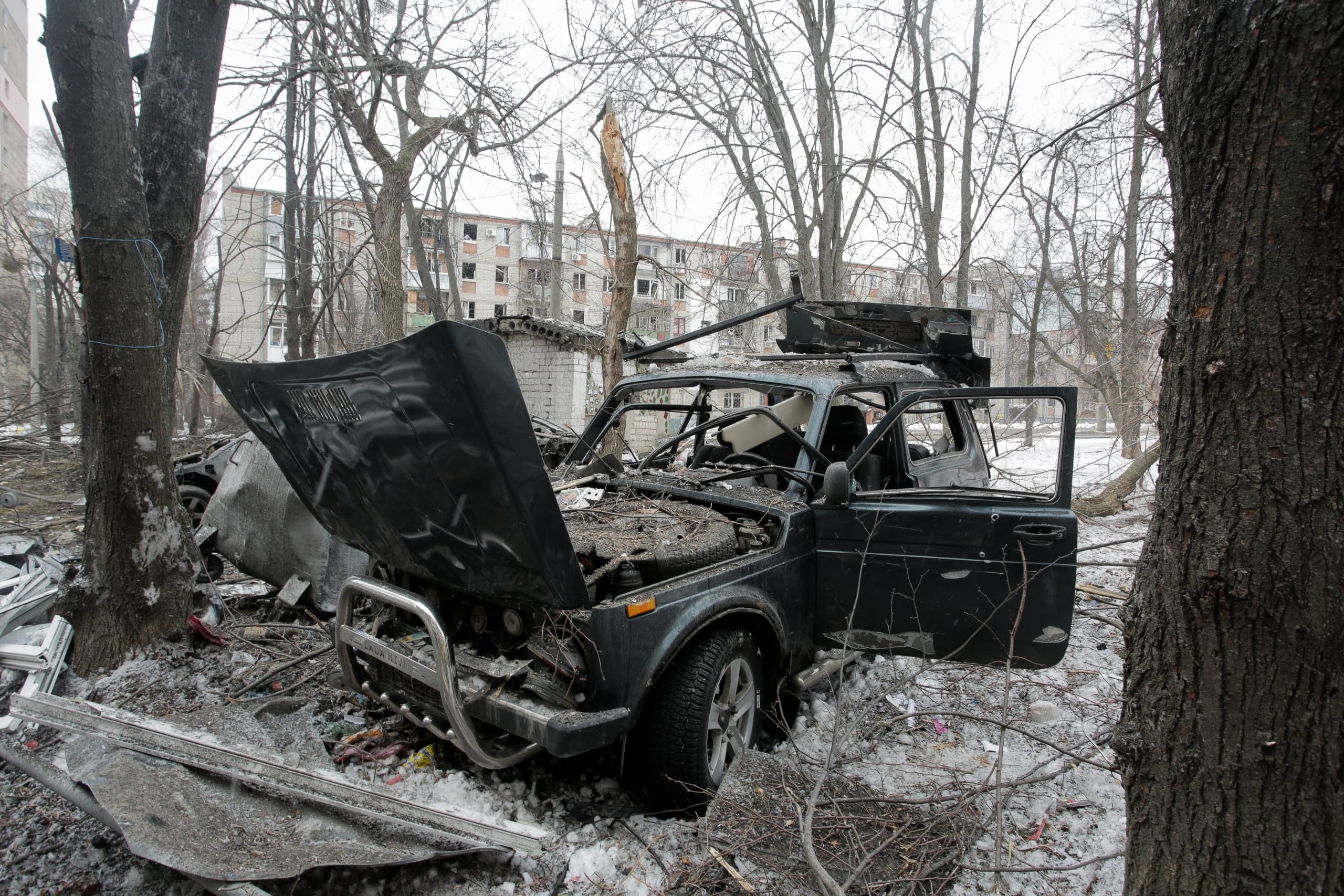 Поврежденный внедорожник Lada Niva с поднятым капотом и разрушенной крышей стоит среди обломков и снега. На заднем плане сквозь голые деревья видны пострадавшие от обстрела многоквартирные дома.
