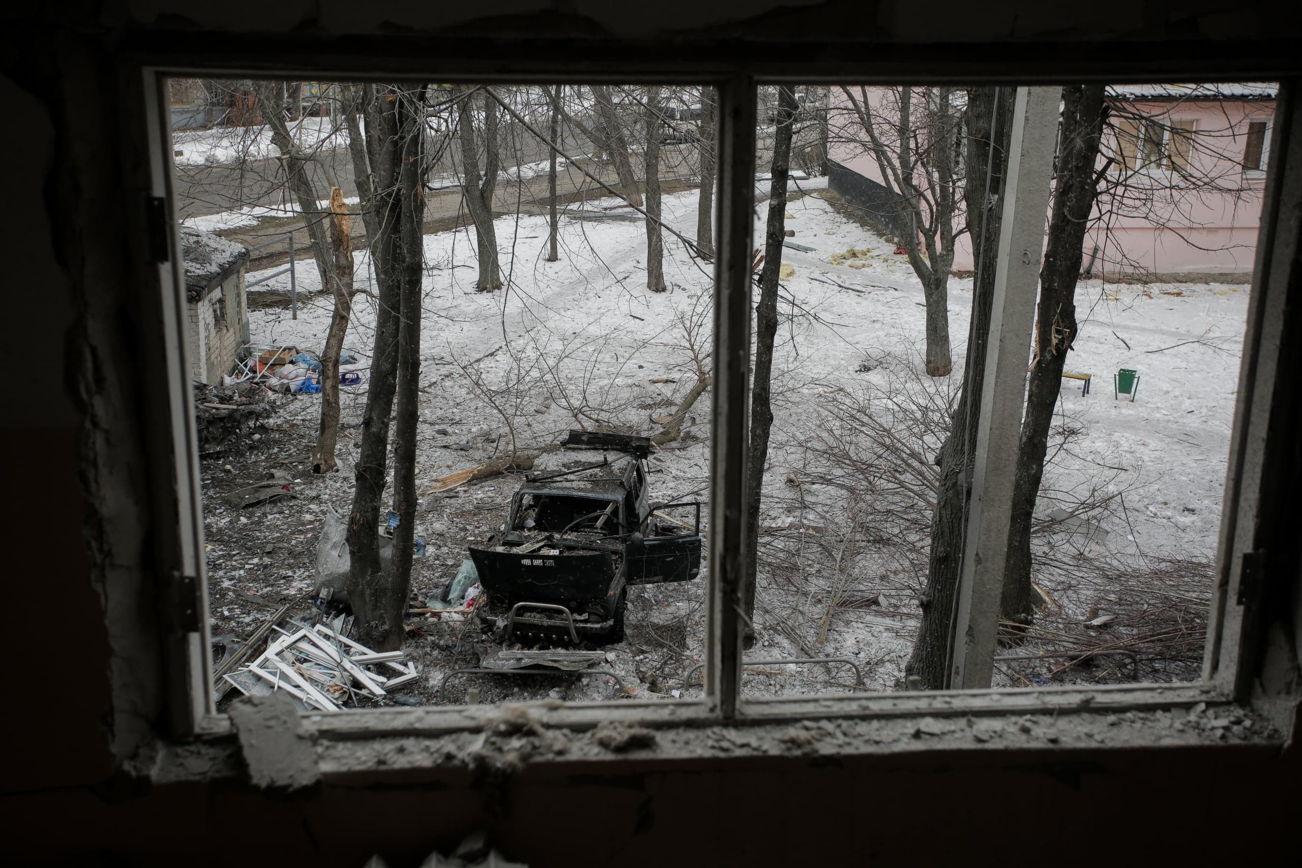 Вид из поврежденного оконного проема на заснеженный двор с обгоревшей машиной, обломками и поваленными деревьями.
