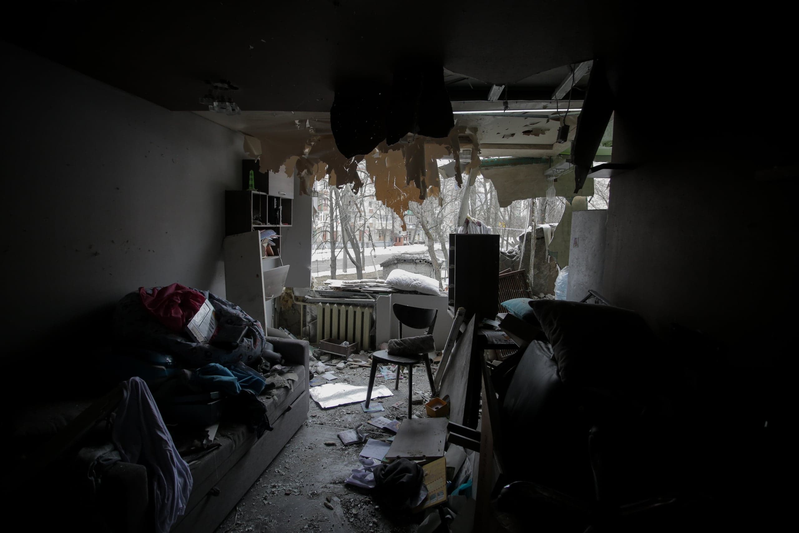 Темный интерьер разрушенной жилой комнаты, с обломками и мусором на полу, с видом на заснеженную улицу через выбитое окно.