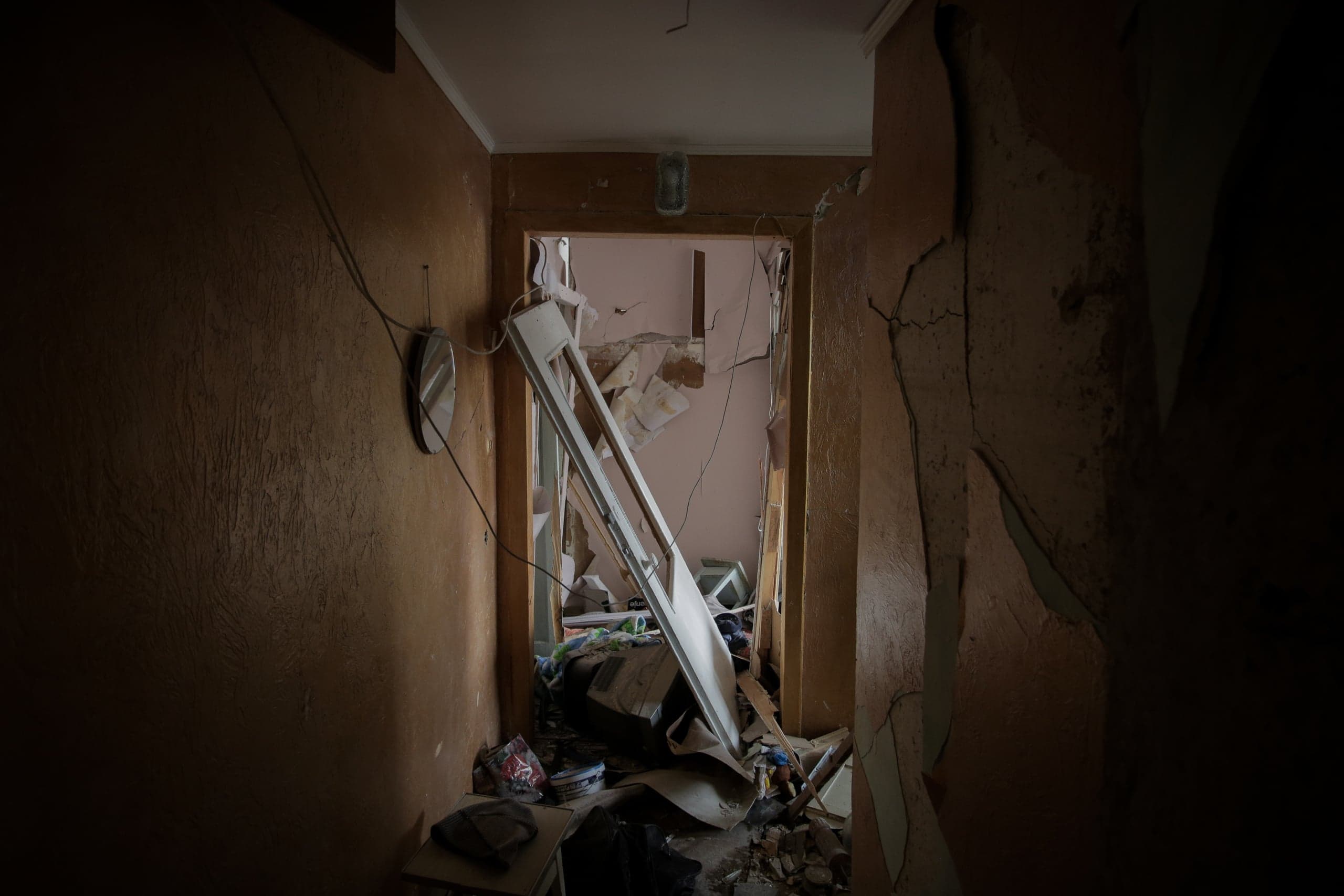 Крупный план сильно поврежденного интерьера: тёмный коридор ведёт в разрушенную комнату, проём которой завален обломками и белой рамой, лежащей по диагонали.