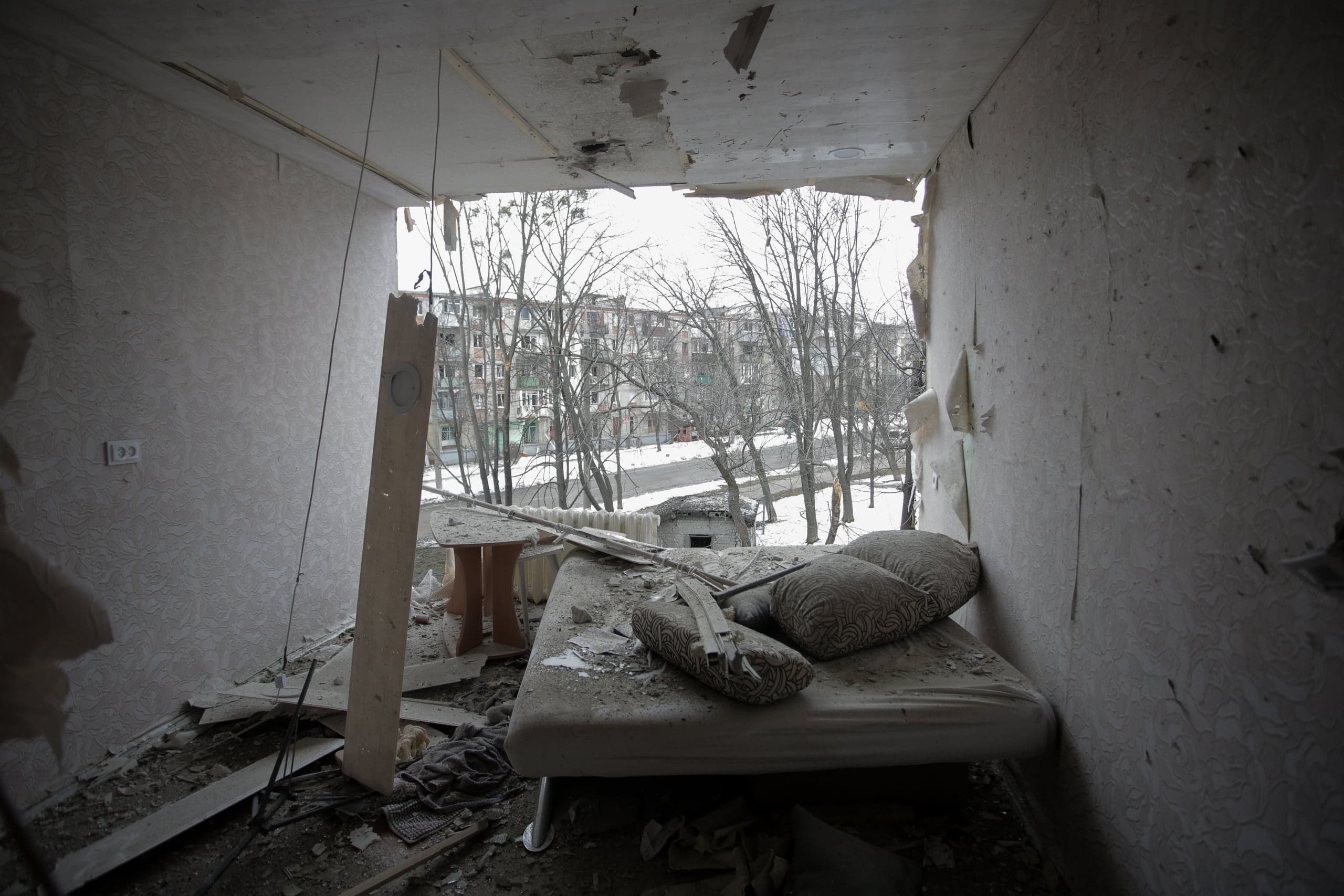 Разрушенная комната, где на месте окна зияет большой проем. Кровать и мебель усыпаны строительным мусором и пылью, за проемом видны голые деревья и многоэтажные жилые дома в зимнем городском пейзаже.