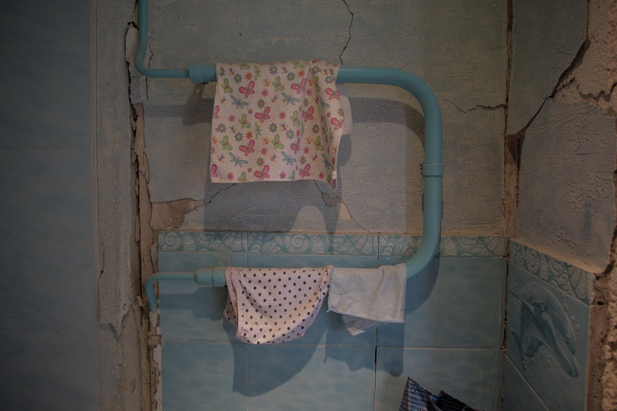 Белье (полотенце с бабочками и трусы в горошек) сушится на голубых трубах на фоне сильно потрескавшейся голубой стены с отвалившейся штукатуркой и декоративной плиткой.