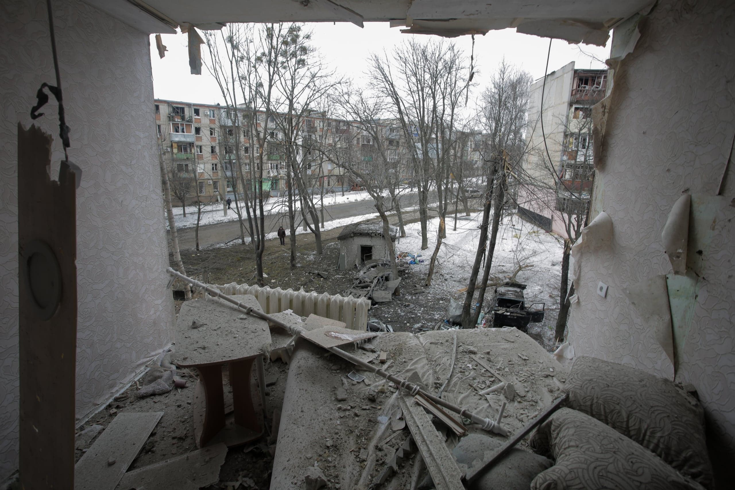 Вид изнутри разрушенной комнаты, заваленной обломками, пылью и поврежденной мебелью. Через выбитый оконный проем виден зимний двор с голыми деревьями, остатками двух сгоревших автомобилей и панельными жилыми домами на заднем плане, многие из которых также повреждены.