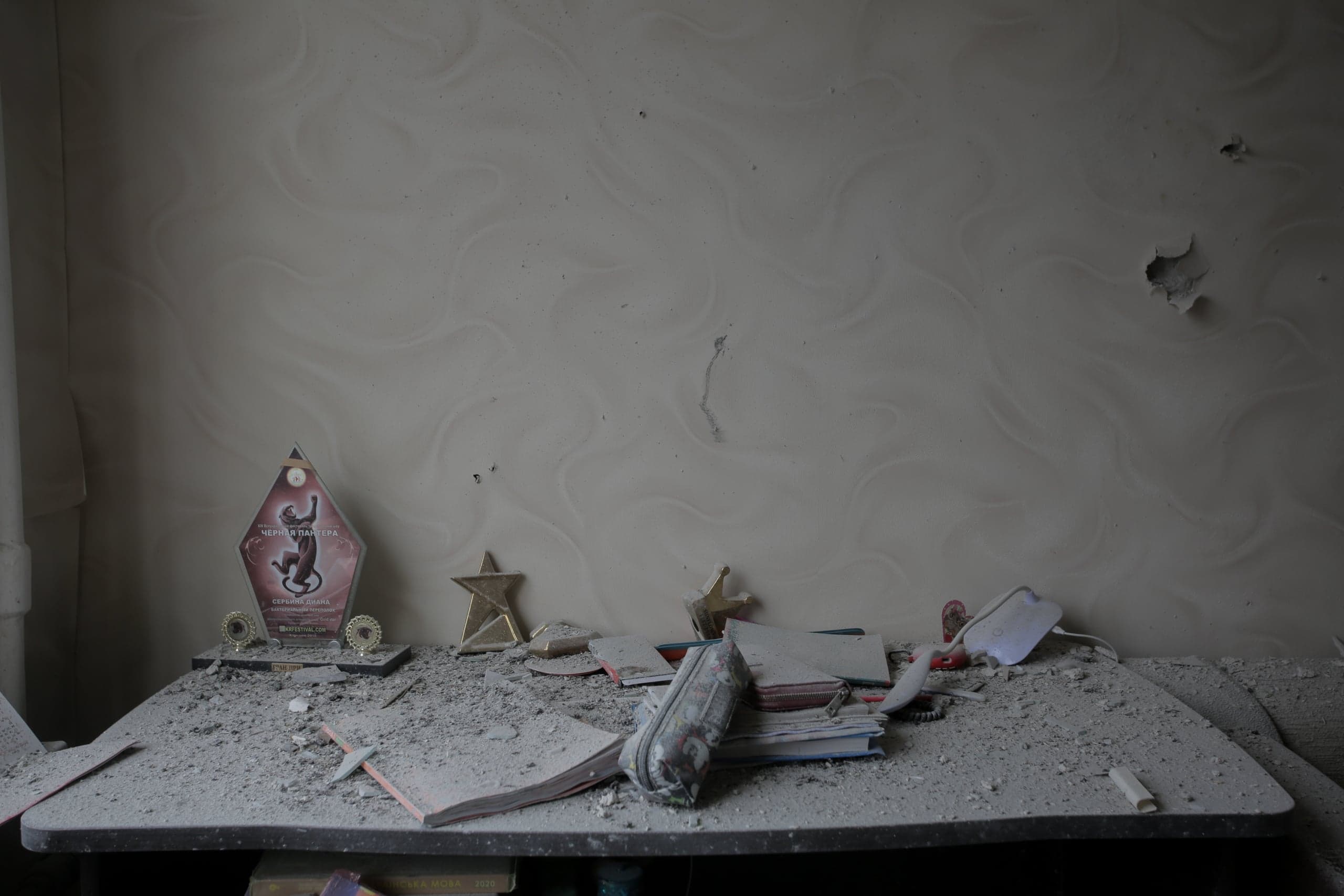 Письменный стол в разрушенной комнате, покрытый толстым слоем серой пыли и обломков. На столе видны школьные тетради, пенал и несколько трофеев. На стене позади стола — следы повреждений.