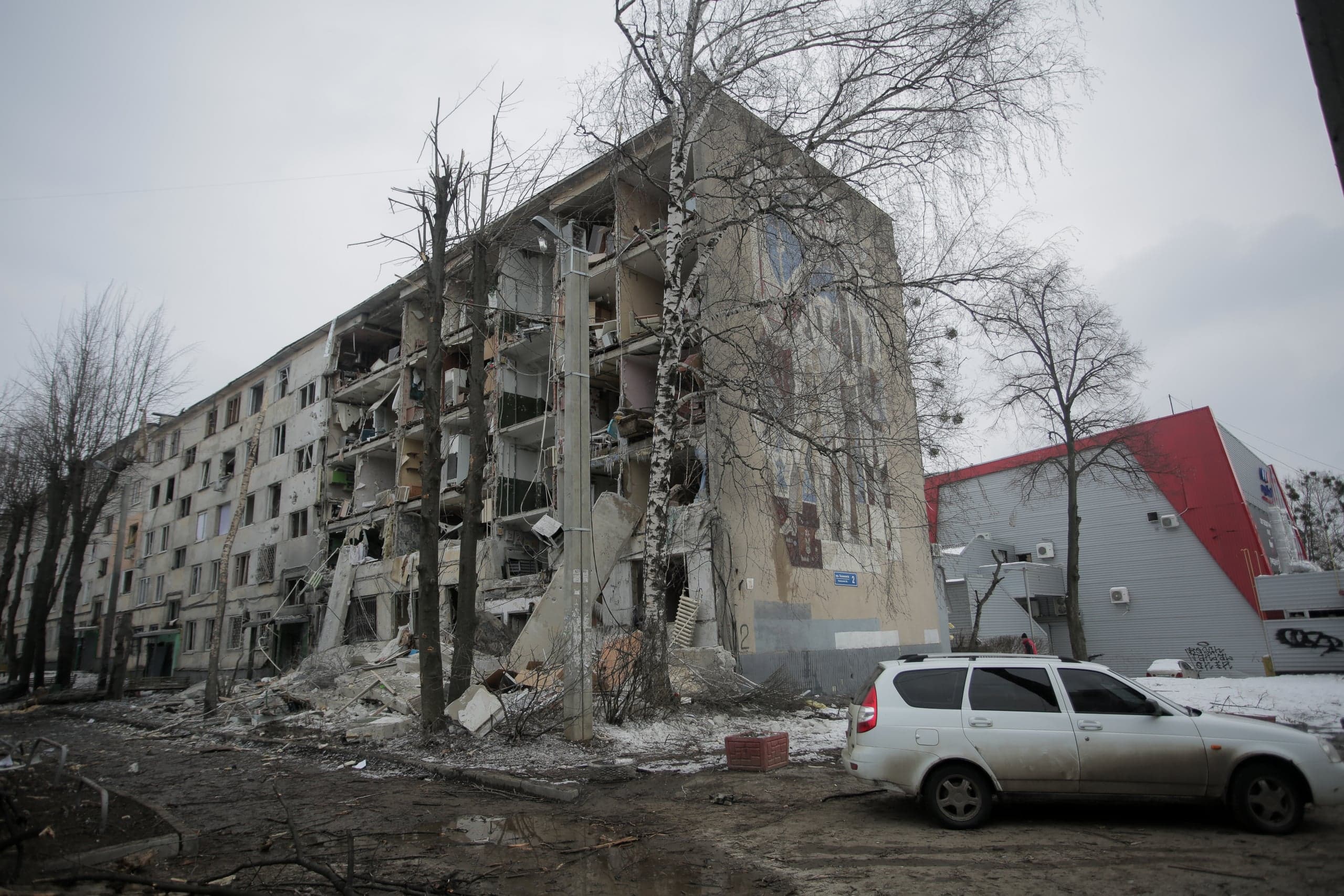 Сильно поврежденный пятиэтажный жилой дом с обрушенной центральной частью и зияющими проемами квартир. Перед зданием лежат завалы и грязь, справа припаркован белый автомобиль-универсал. Небо пасмурное.