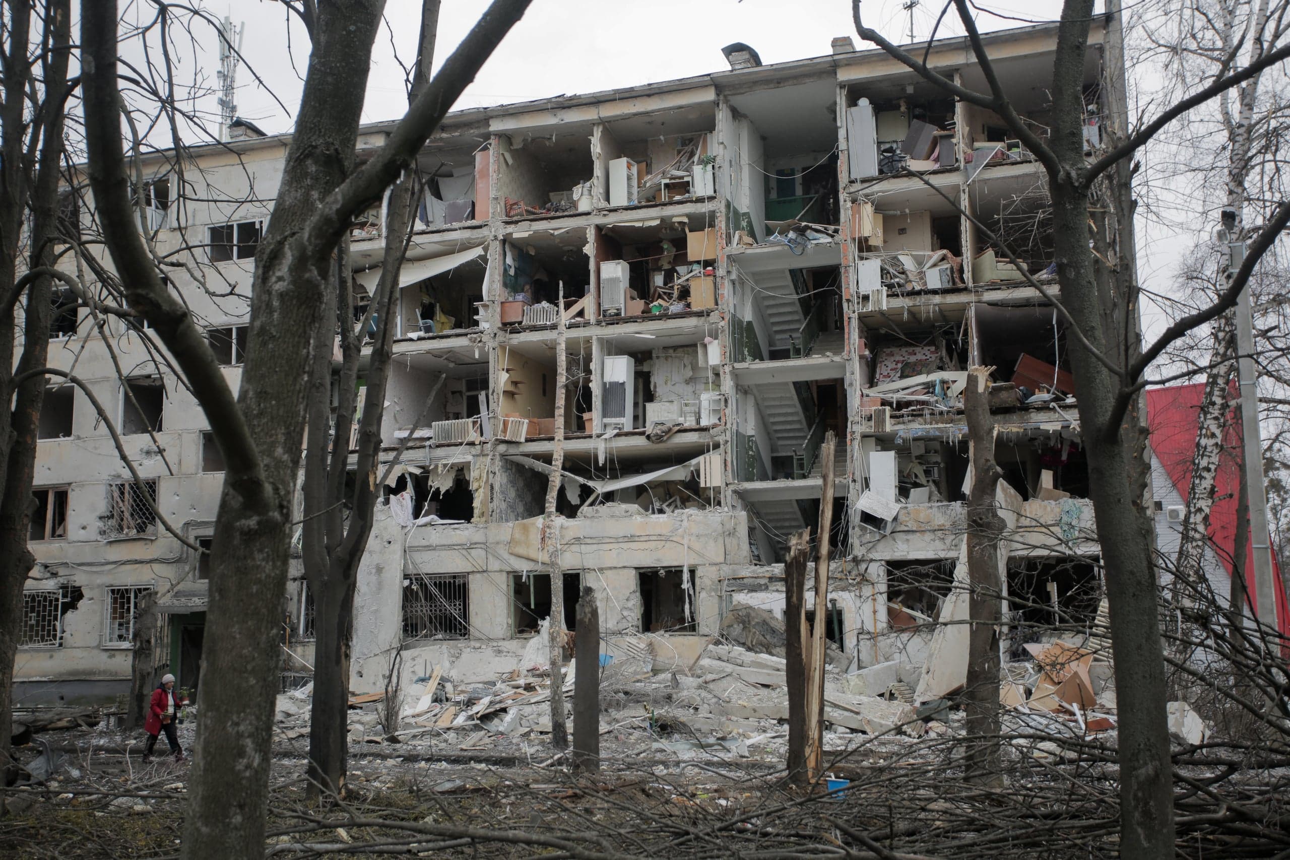 Сильно разрушенный пятиэтажный жилой дом с обрушенной центральной секцией, обнажающей лестничные пролеты и интерьеры квартир. Огромное количество строительного мусора и обломков перед зданием. Снимок сделан сквозь голые ветви деревьев; в левой части кадра виден человек в красной куртке.