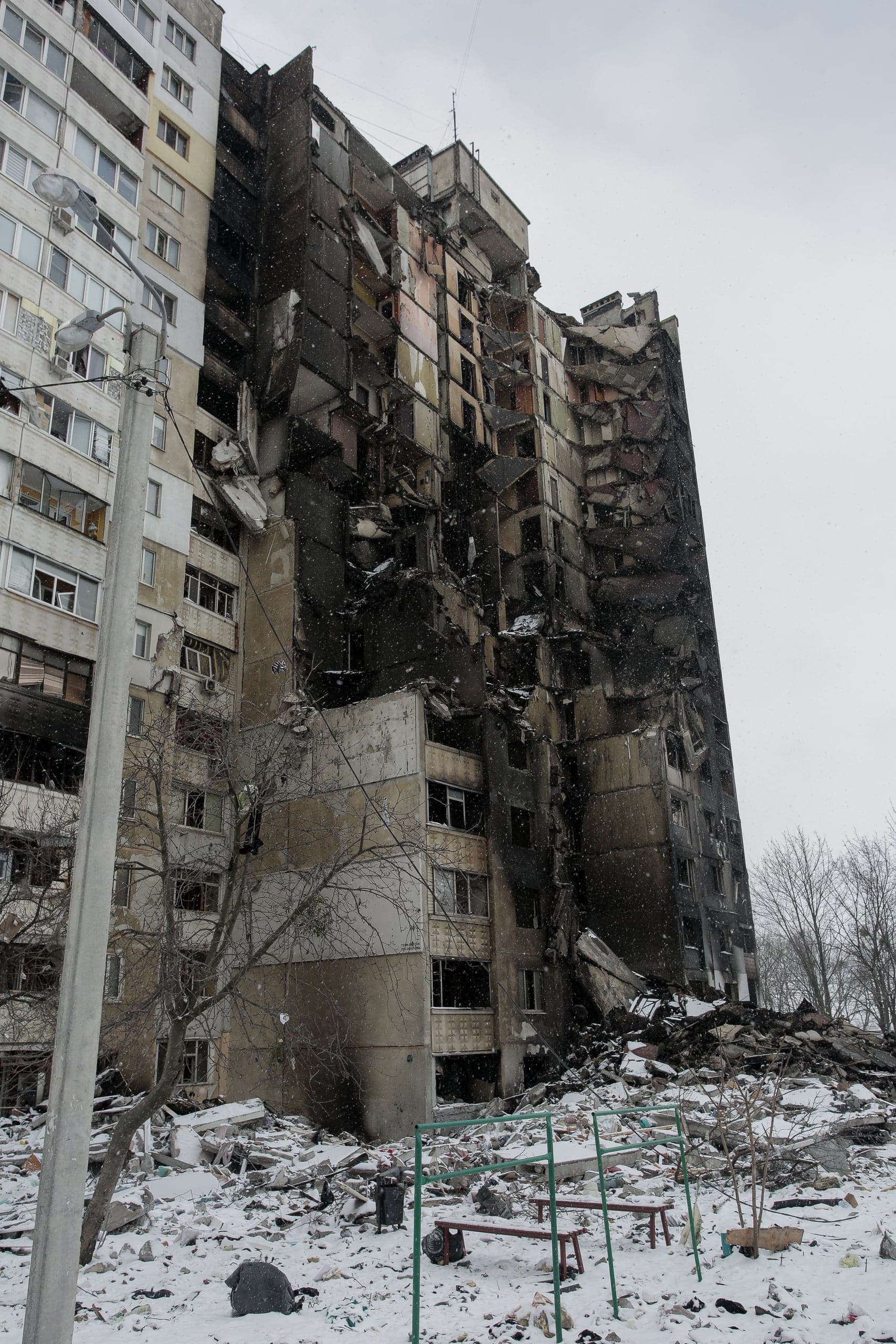 Сильно поврежденный, почерневший многоквартирный дом среди снега и руин.