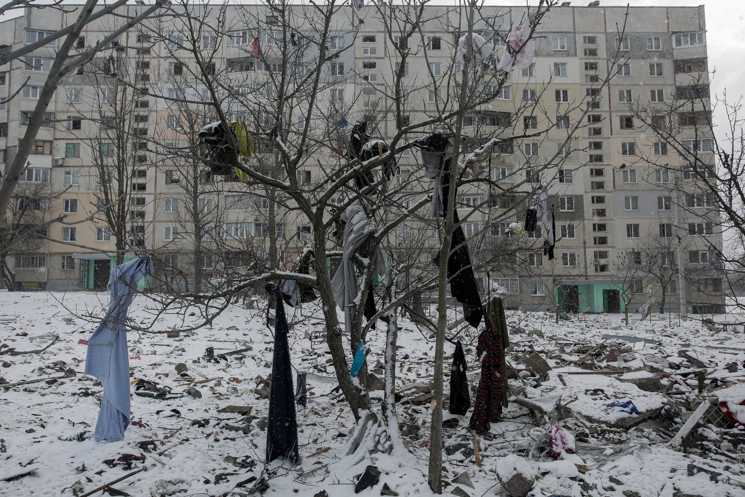Снежный пейзаж с разрушениями: деревья, увешанные одеждой, перед многоквартирным домом, двор покрыт мусором и обломками.