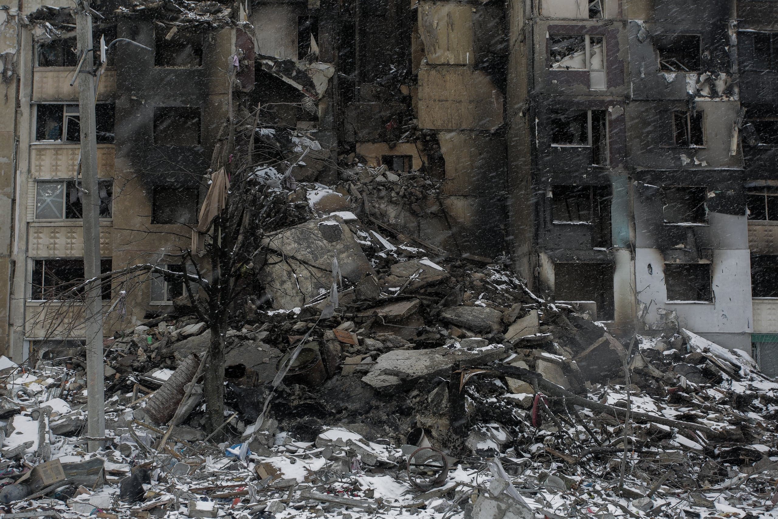 Разрушенное многоэтажное здание, груда обломков на снегу под снегопадом. Фасад обуглен.