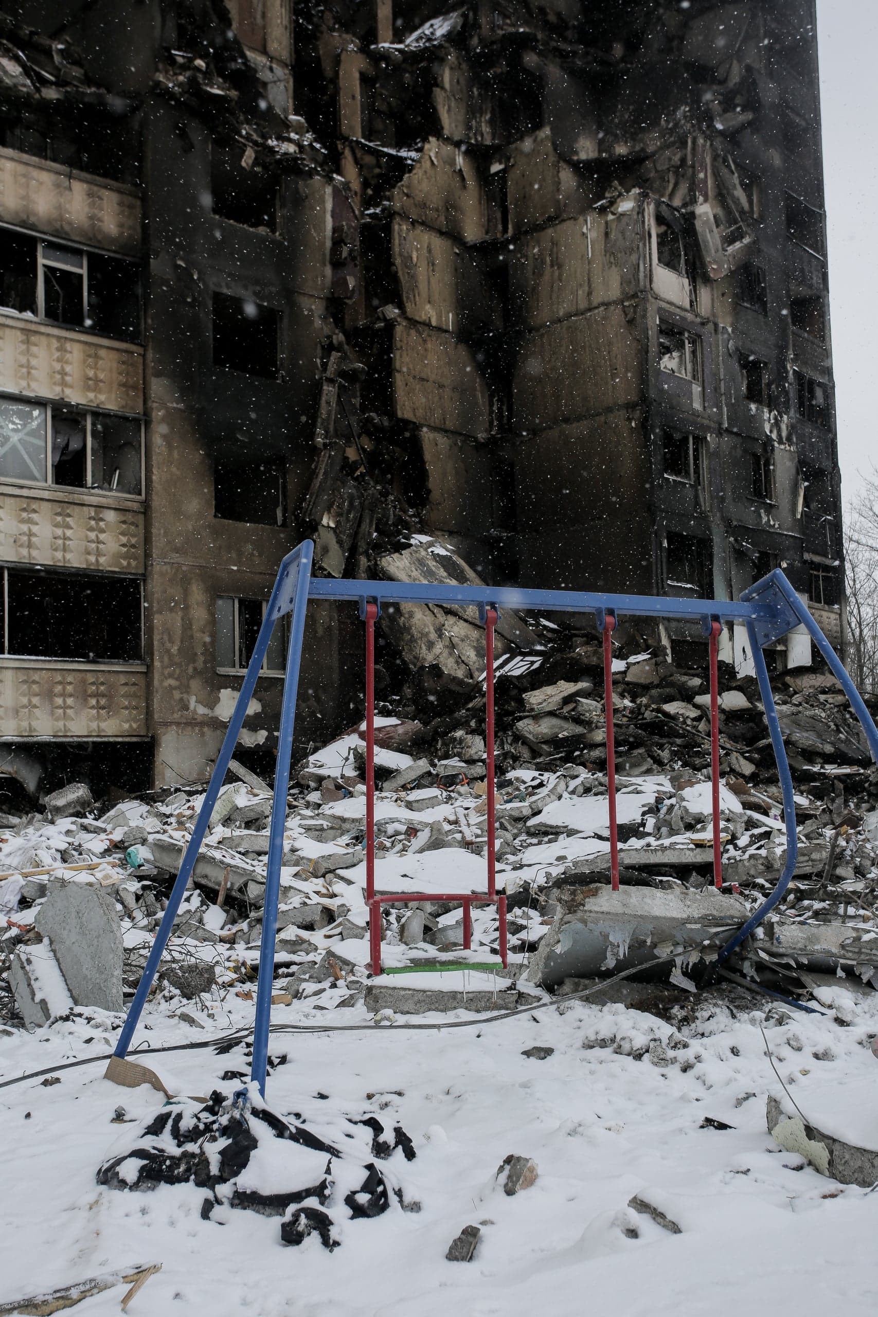 Сломанные качели на снегу перед разрушенным и обугленным многоэтажным домом.