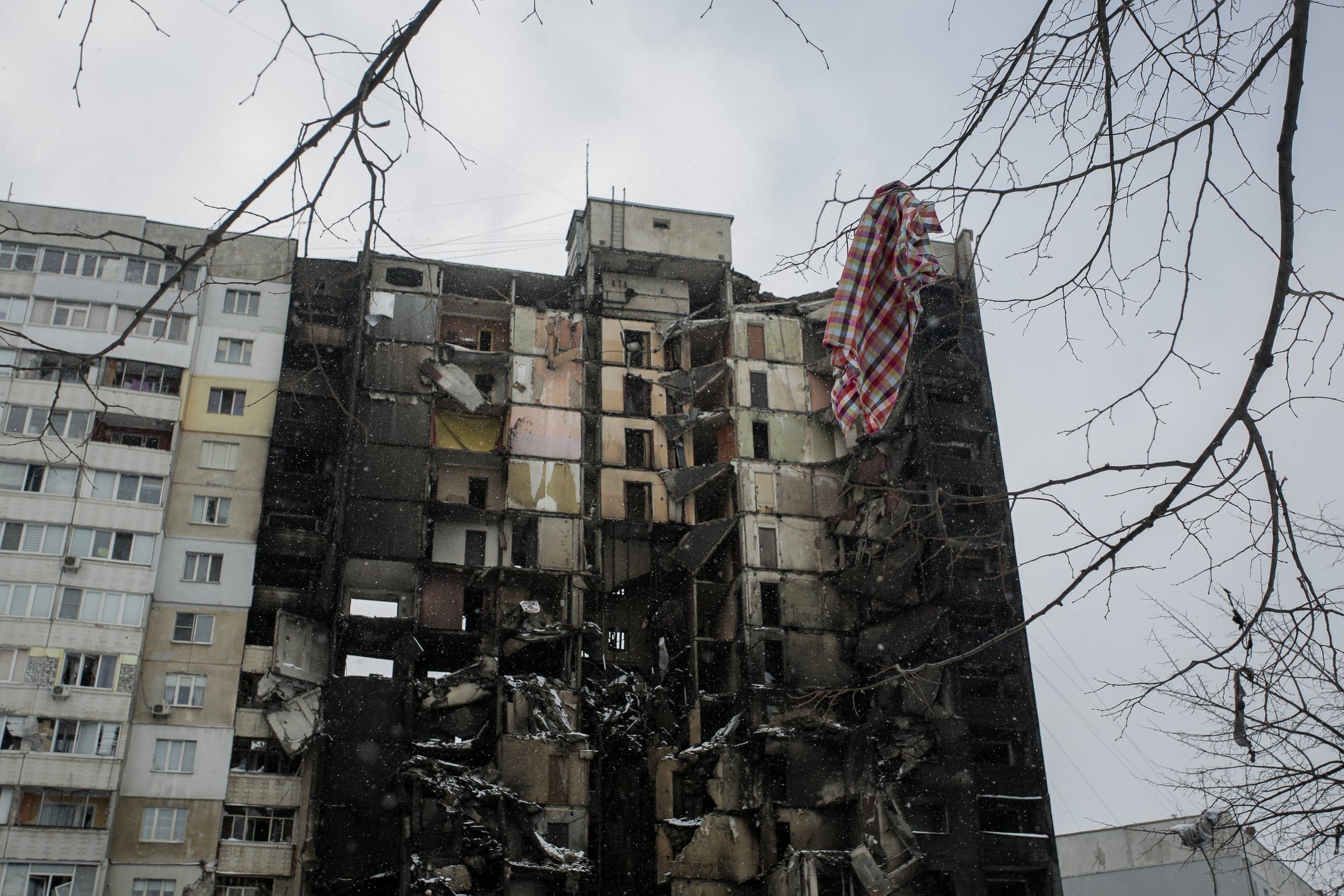 Поврежденный и обгоревший многоэтажный жилой дом с выбитыми окнами и разрушениями, рядом целая секция; на крыше свисает клетчатая ткань.