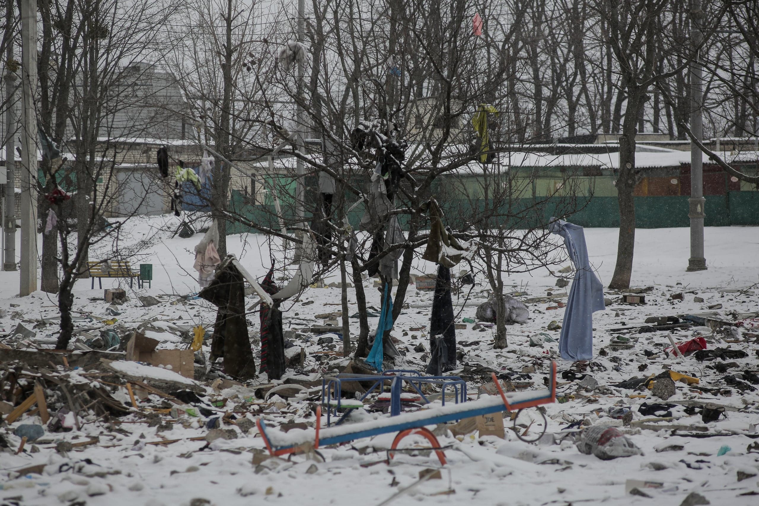 Снежный пейзаж разрушенного двора. На земле обломки и мусор, на переднем плане детские качели-балансир. Разбросанная одежда висит на голых ветвях деревьев.