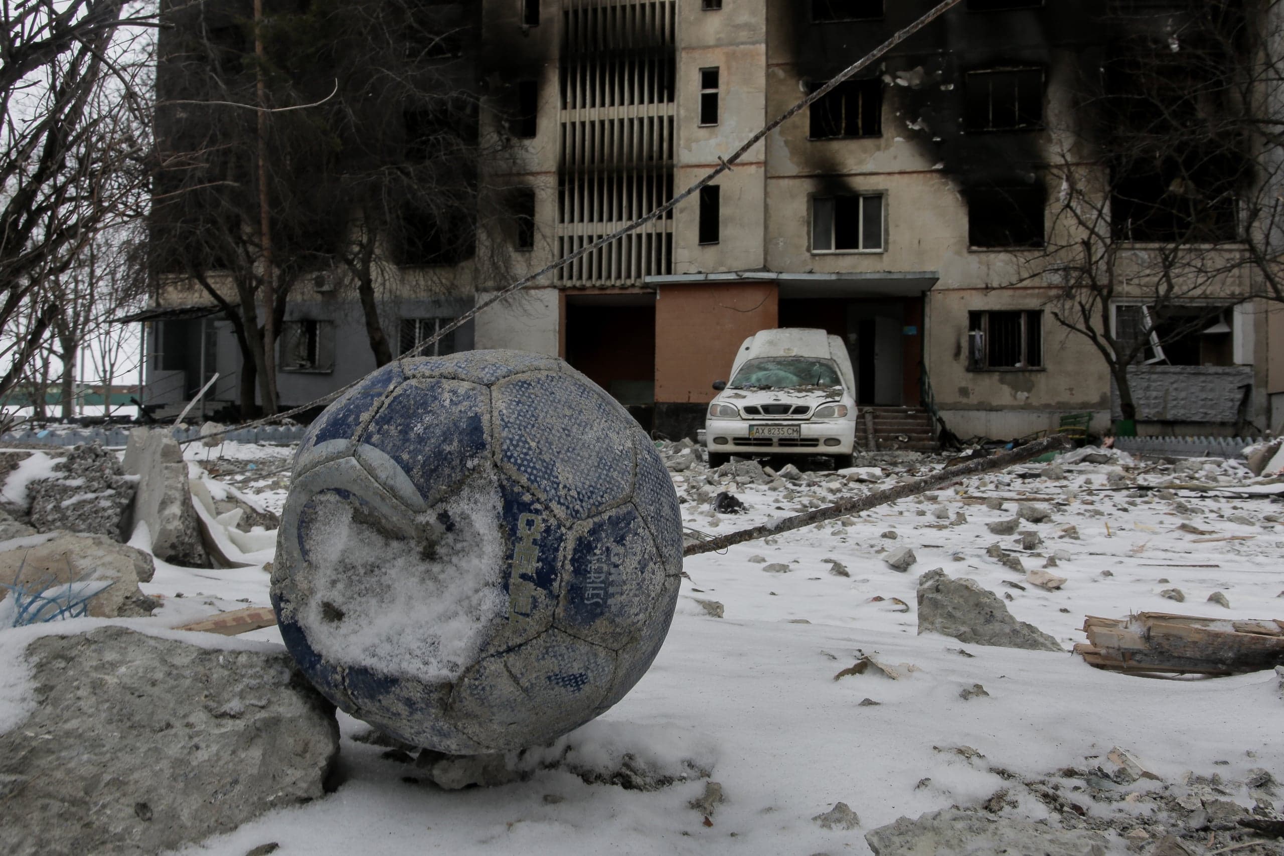 Изношенный футбольный мяч среди снега и руин на фоне поврежденного огнем жилого дома и белого фургона.