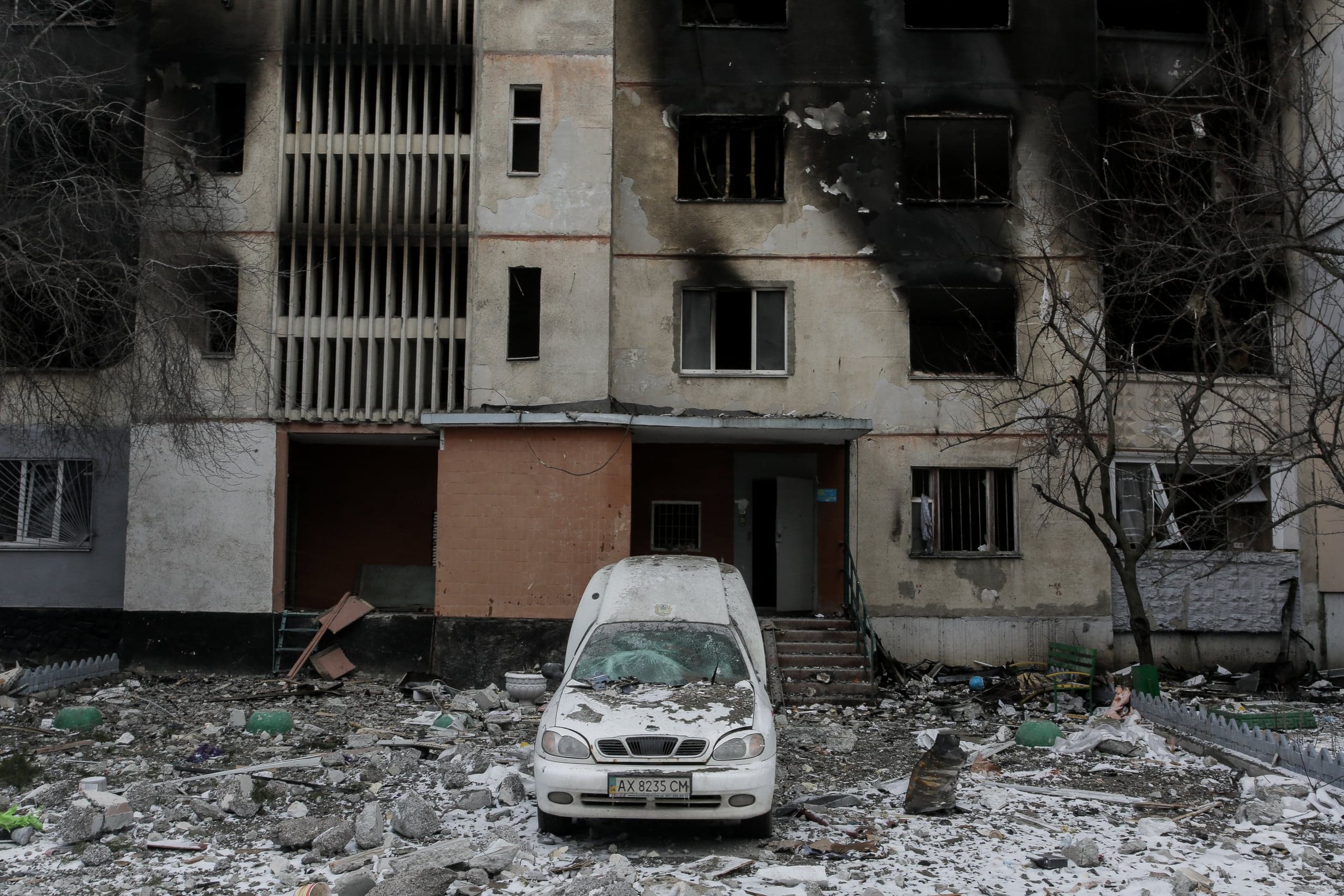Разрушенный фасад жилого здания после обстрела, следы копоти. Среди обломков стоит белая машина с разбитым стеклом, номер "AX 8235 CM".