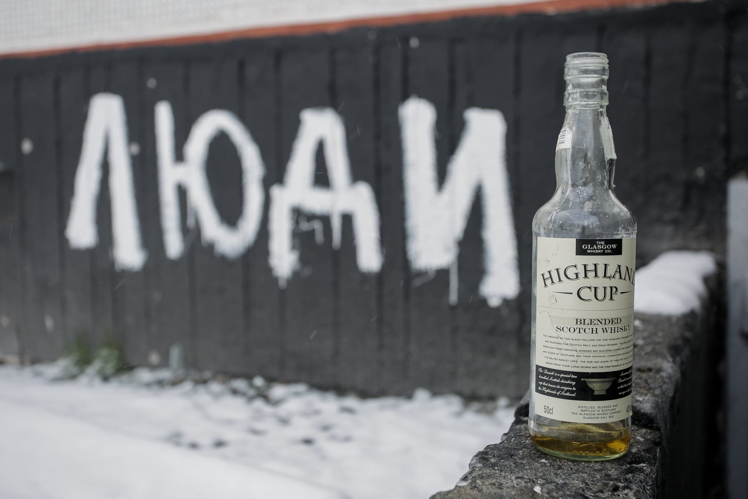 Полупустая бутылка виски "Highland Cup" на фоне стены с белым граффити "ЛЮДИ".