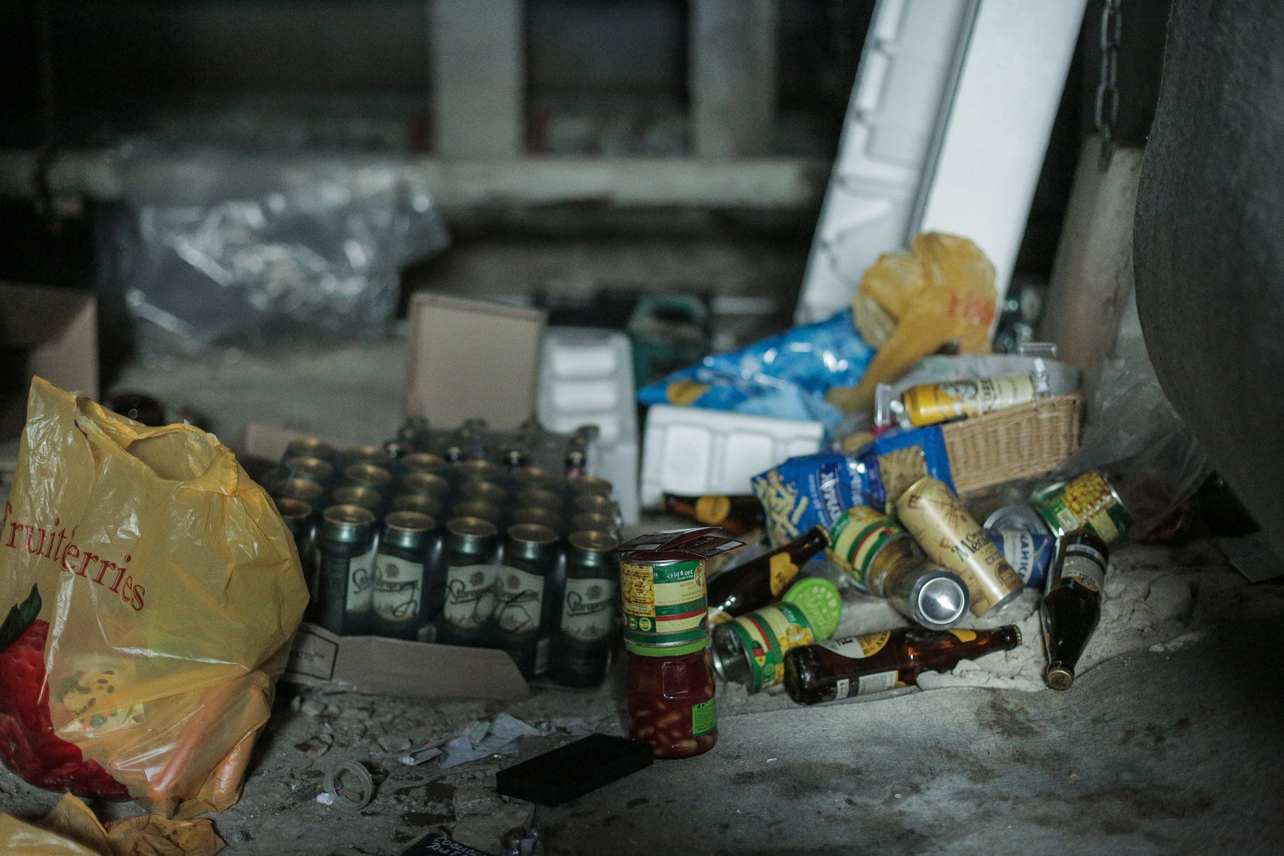Складское помещение, заваленное мусором и припасами: ящик банок, желтый пакет с надписью "fruiterries", консервы и разбросанные пустые бутылки на грязном полу.