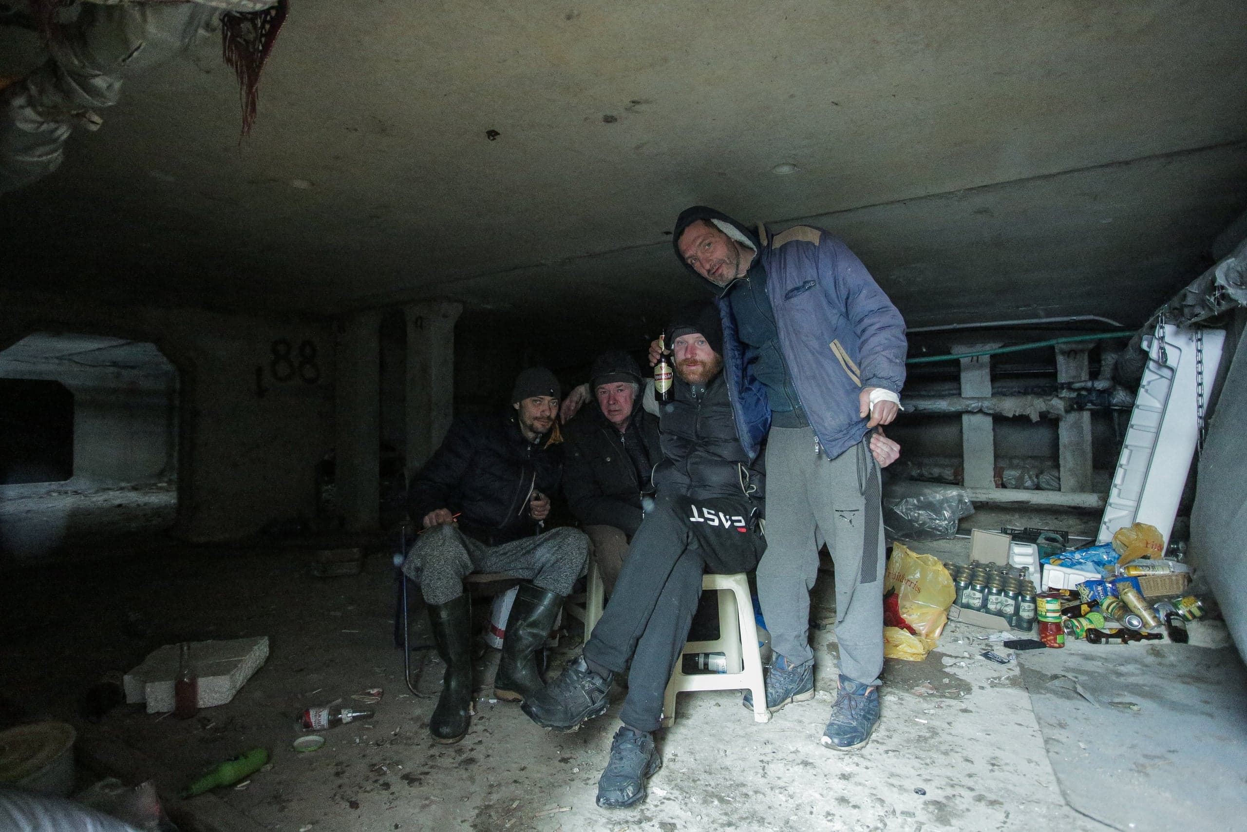 Четверо мужчин сидят или стоят в грязном, темном, низком подвальном помещении, окруженные мусором и бутылками. На стене виден граффити "88".