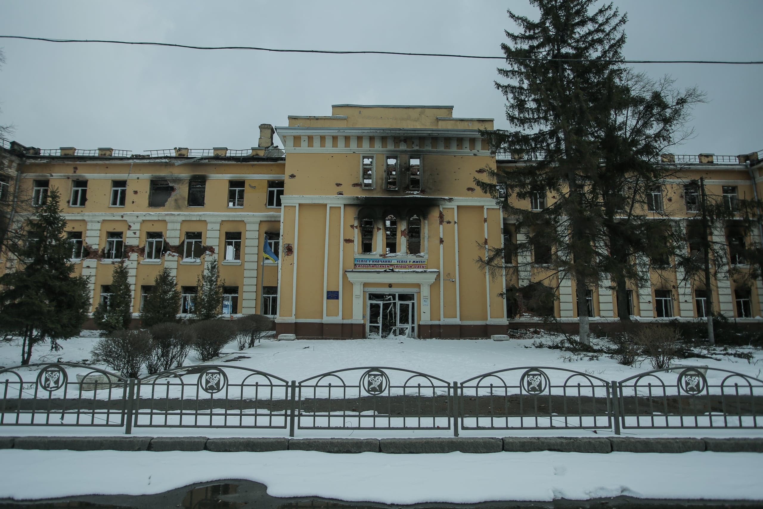 Сильно поврежденное обстрелами желтое здание со следами гари над входом. Зимний пейзаж.