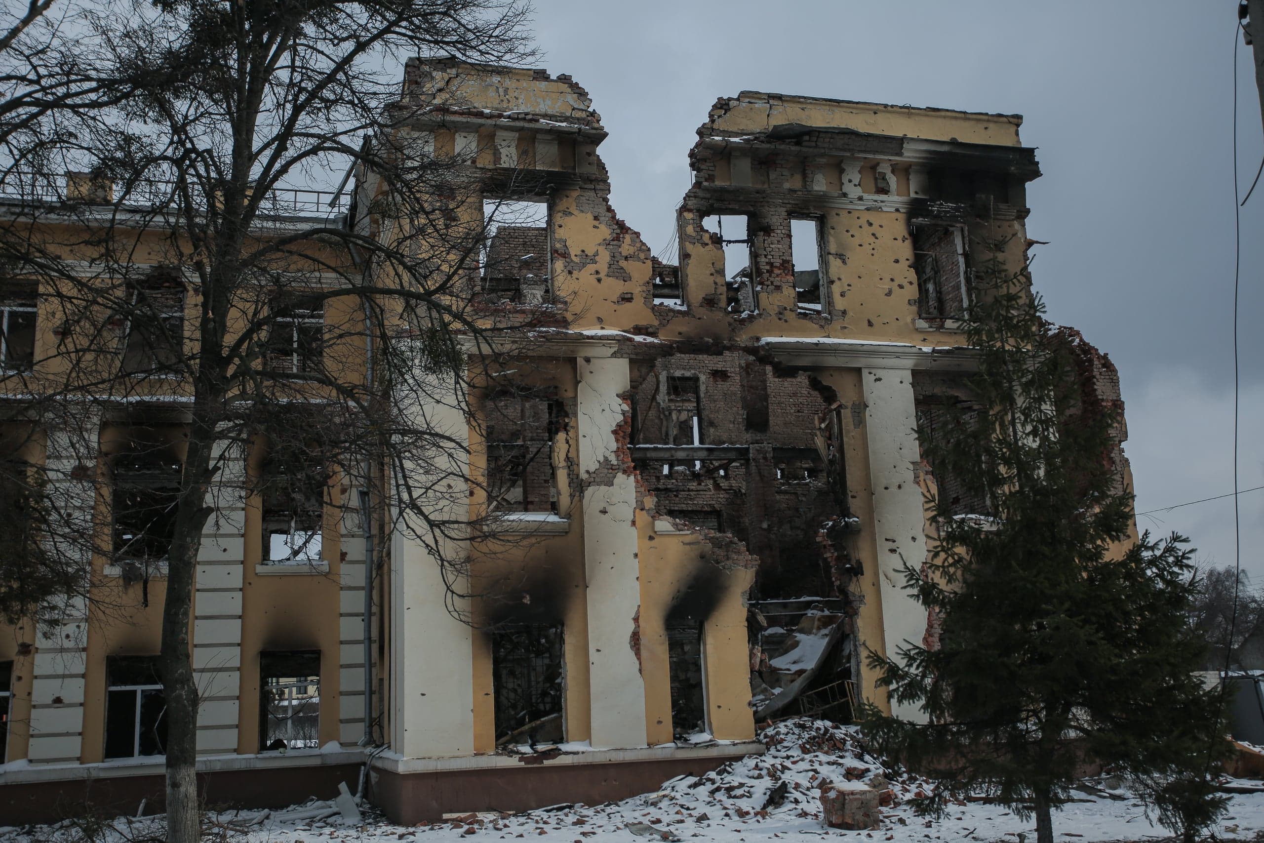 Сильно поврежденное обстрелом желтое здание с обрушенной центральной частью на фоне зимнего пейзажа.