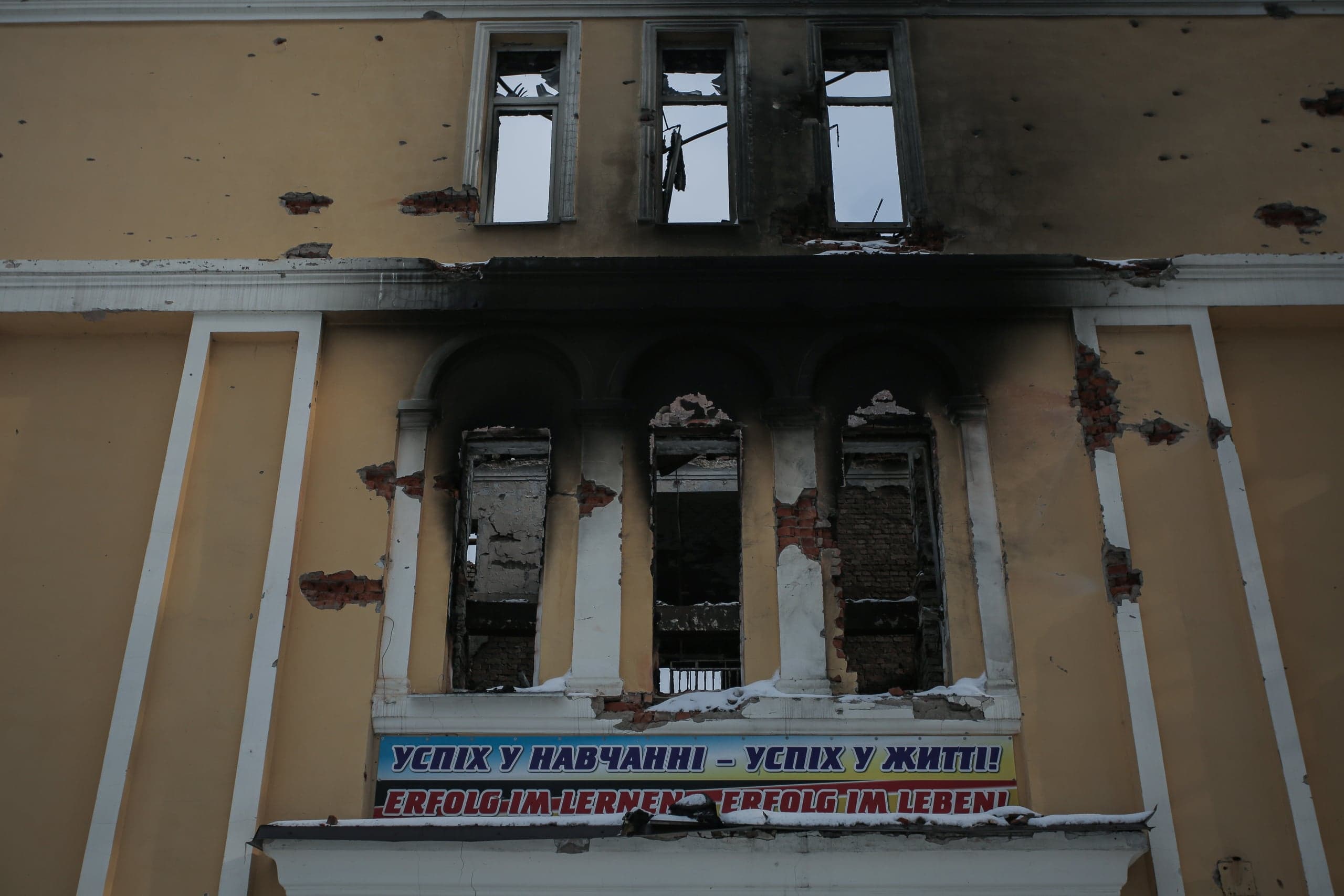 Поврежденный обстрелом фасад здания с выбитыми окнами, следами пожара и баннером: "УСПІХ У НАВЧАННІ – УСПІХ У ЖИТТІ! ERFOLG IM LERNEN ERFOLG IM LEBEN!".
