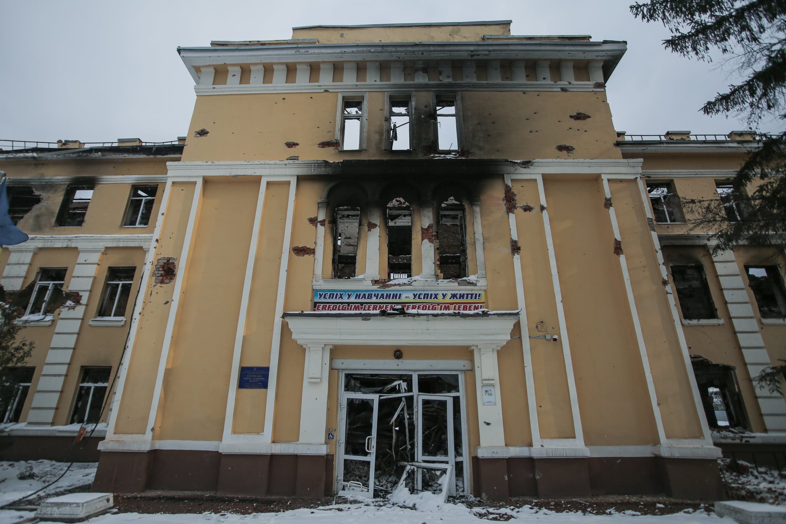 Поврежденное, обугленное здание с выбитыми окнами, следами попаданий и разрушенным входом. Над входом баннер: "УСПІХ У НАВЧАННІ – УСПІХ У ЖИТТІ! ERFOLG IM LERNEN – ERFOLG IM LEBEN!". Снег на земле.