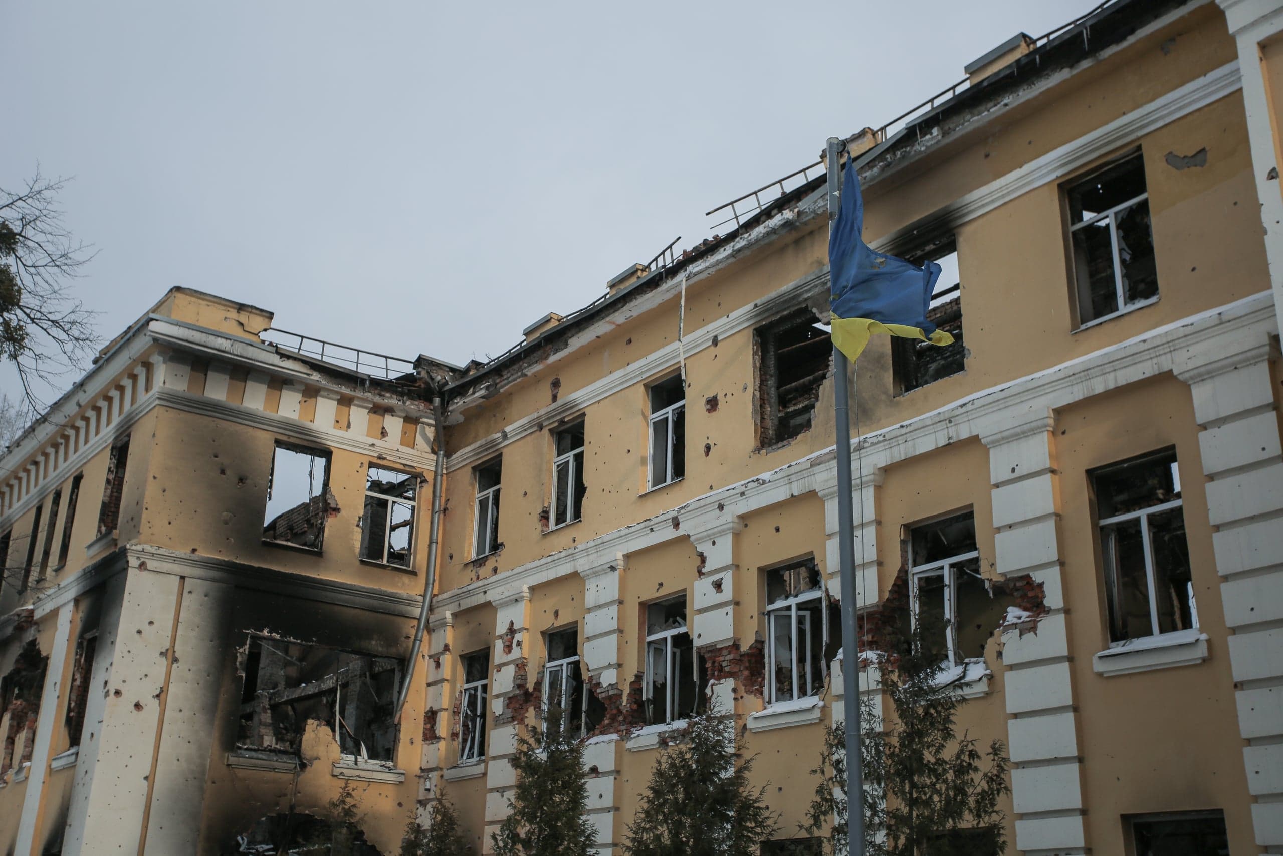 Поврежденное, обгоревшее здание с множеством следов от обстрелов, выбитыми окнами и порванным флагом Украины на флагштоке.