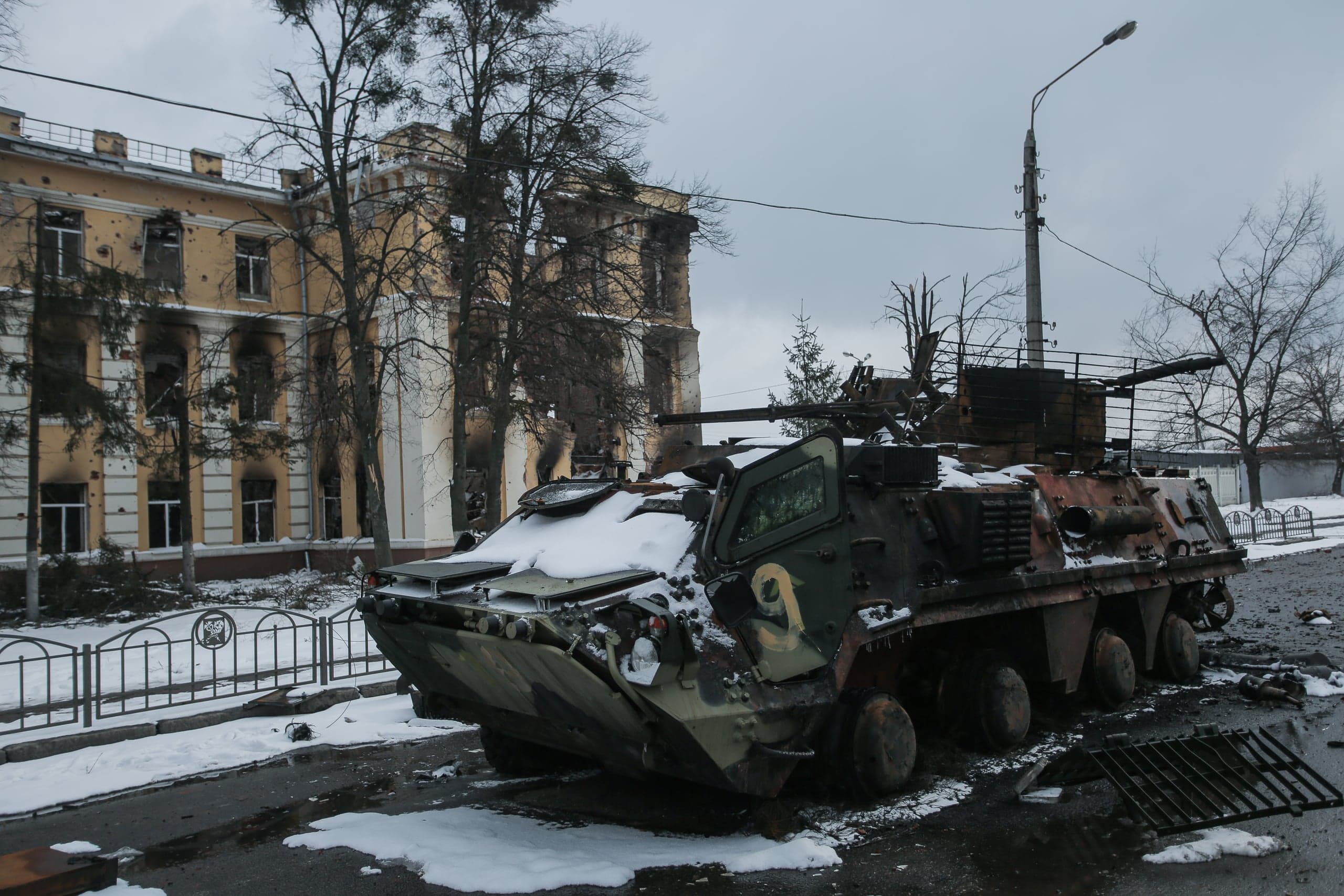 Подбитый, частично заснеженный бронетранспортер на фоне поврежденного, обгоревшего здания.